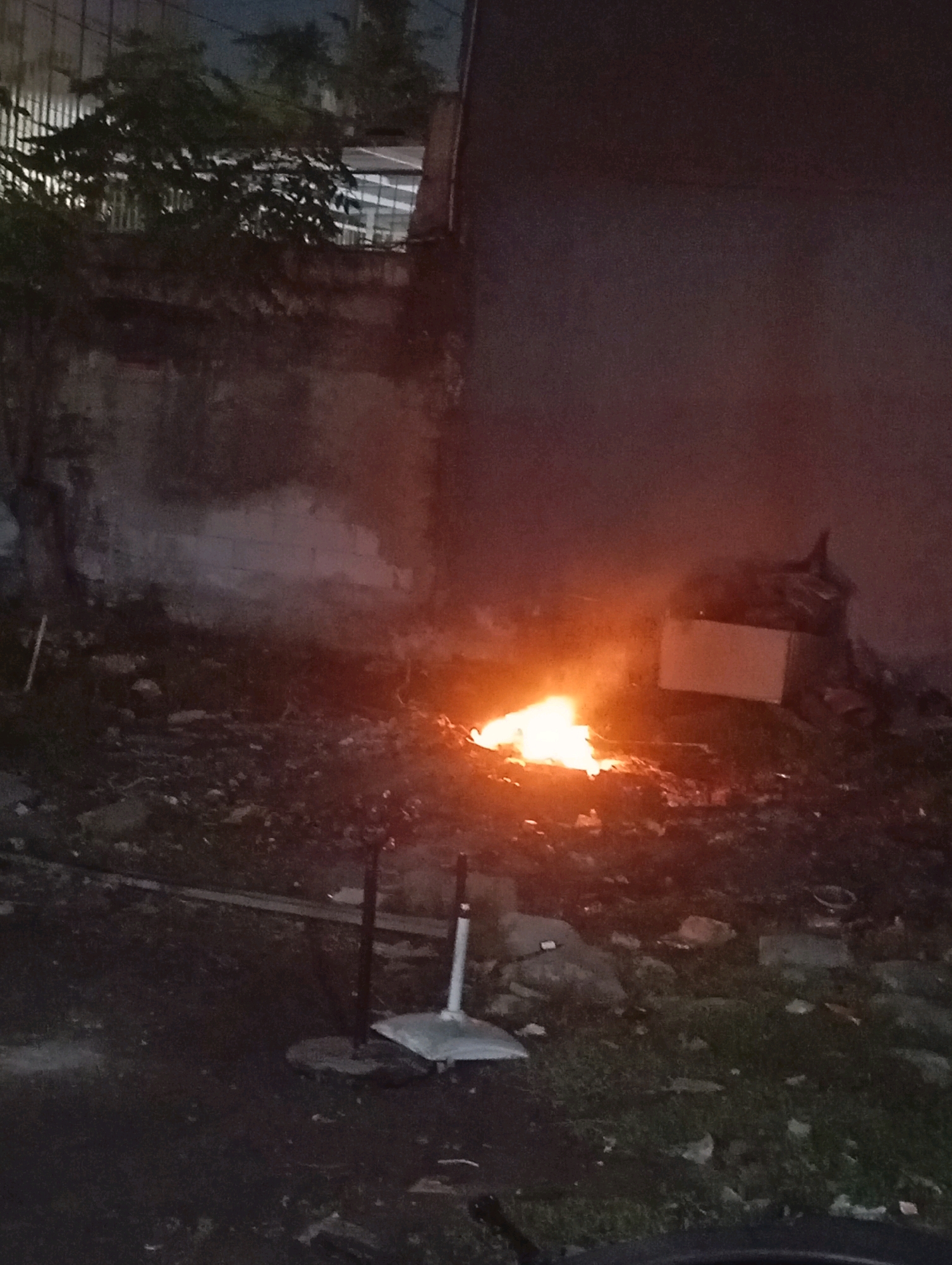 A nighttime outdoor scene depicts a small fire burning in a pile of debris next to a textured, weathered wall. The fire emits a bright orange glow and light smoke. In the foreground, a dark asphalt surface is visible with some scattered debris and patches of grass. Two dark poles with white attachments stand in the middle ground. To the right of the fire, a light-colored, rectangular object resembling a piece of furniture or appliance sits amongst the debris, with a dark silhouette of an object or person atop it. The background shows more debris and a dark, indistinct structure with some foliage. The overall atmosphere is dark and somewhat ominous due to the fire and the nighttime setting.