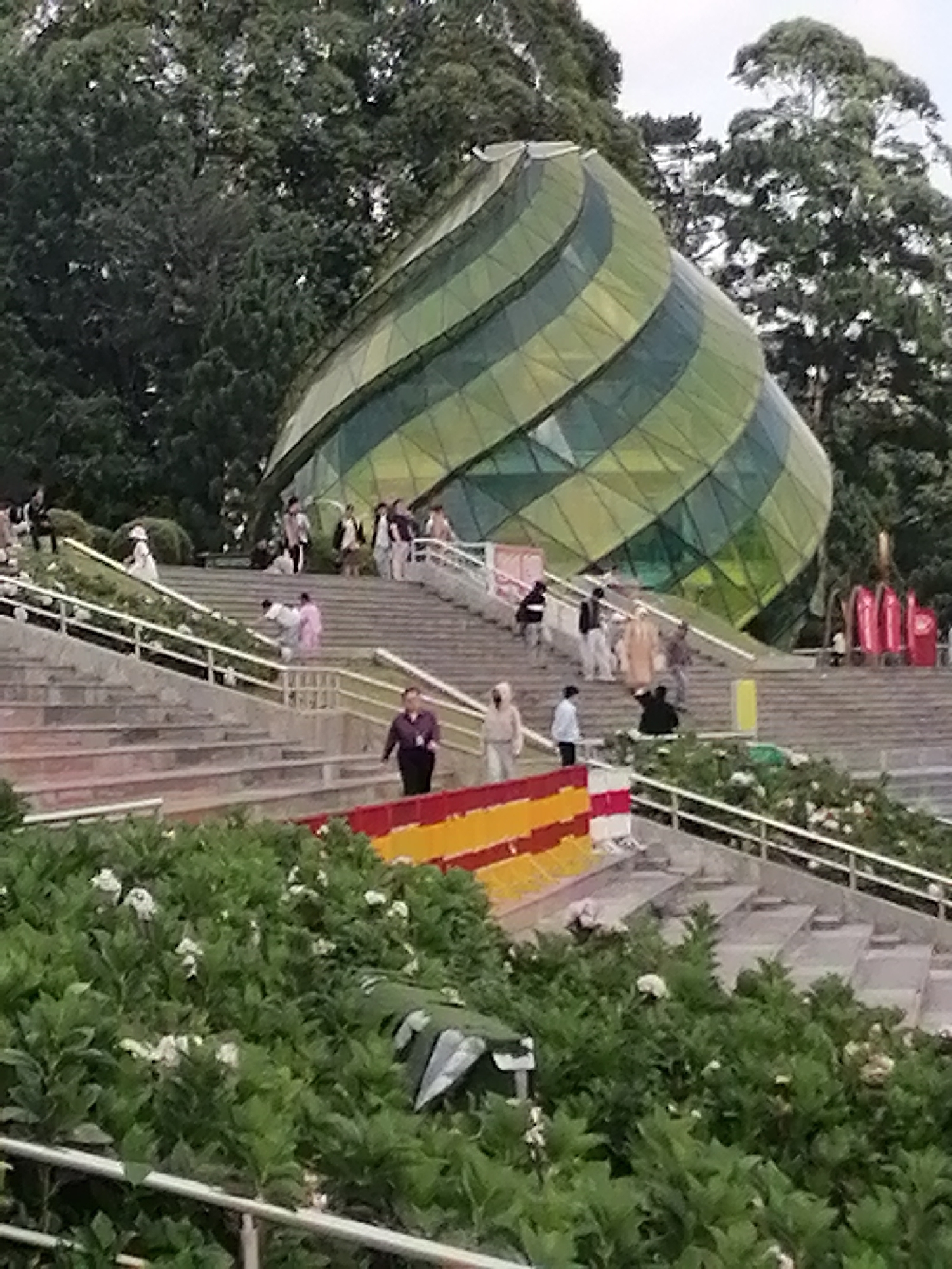 The image depicts a wide shot of the entrance to the Da Lat Flower Park in Da Lat, Vietnam. A striking, spiral-shaped glass structure with green and yellow panels dominates the upper right portion of the frame, set against a backdrop of dense green trees. In front of the structure, a broad set of concrete stairs ascends, lined with white metal railings.
Scattered across the stairs are numerous individuals. On the lower steps, a person in a purple shirt walks down. Further up, a group of people are gathered, some in traditional Vietnamese attire. Several individuals are walking up and down the stairs, while others appear to be observing the surroundings.
The foreground is filled with lush green foliage and white flowers, suggesting a well-maintained garden. Along the edge of the stairs, a horizontal banner displays bold red, yellow, and white stripes. The overall scene conveys a public gathering space, likely a tourist attraction, with a blend of modern architecture and natural elements. The sky is overcast, suggesting a cloudy day.