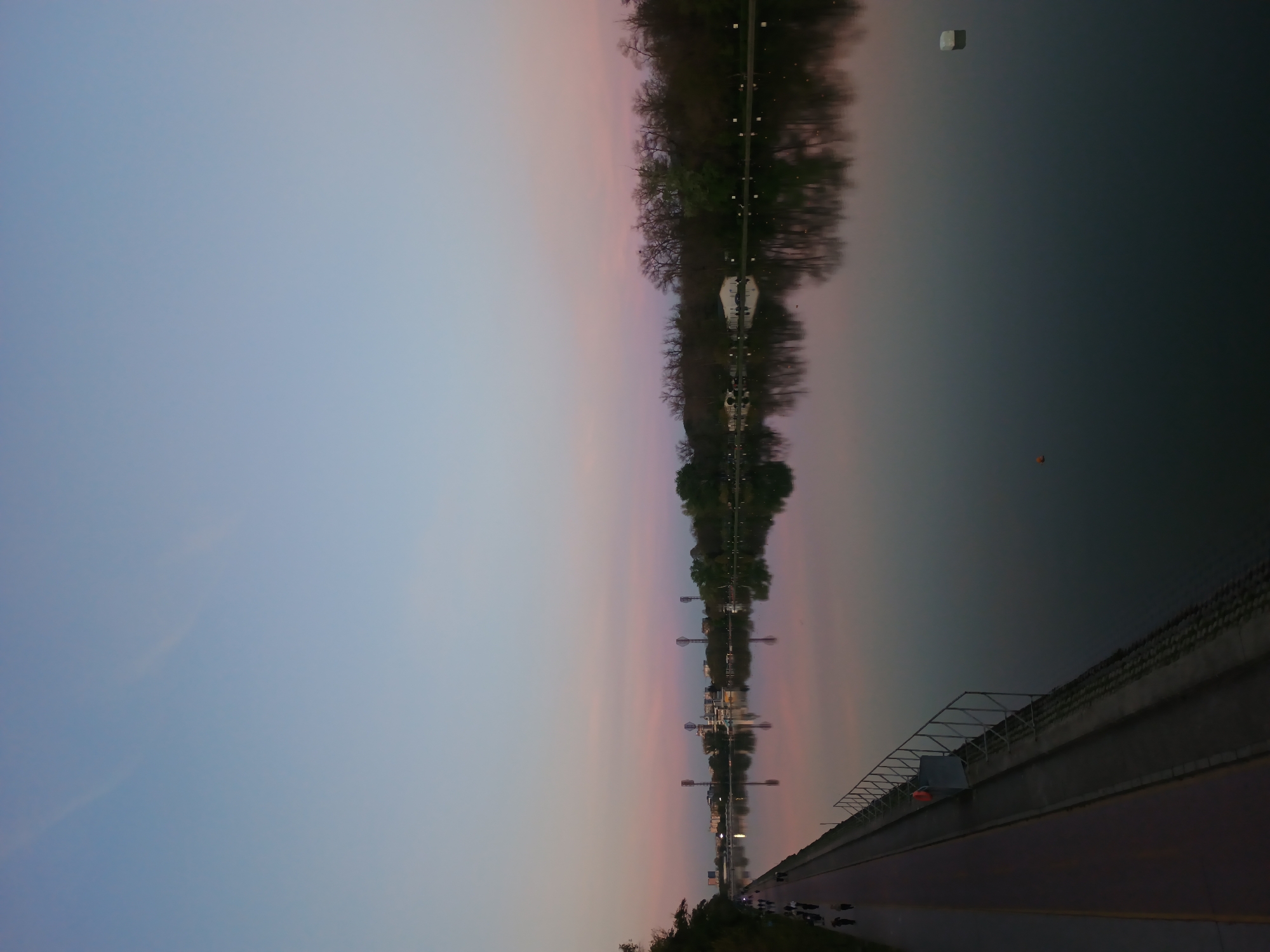 The image captures a serene landscape of a wide river or regatta course at dusk or dawn, likely the Regatta Course in Plovdiv, Bulgaria. The sky, across the top, displays a soft gradient of blue transitioning into gentle pink and orange, characteristic of twilight.
The calm water, occupying the middle ground, acts as a mirror, reflecting the colorful sky and the tree-lined opposite bank. Silhouetted trees, some appearing bare, and faint architectural structures are visible on the far bank, their reflections extending into the water. Several light pylons or decorative posts also dot the distant shoreline, casting elongated reflections. Floating on the water are a white rectangular object and a smaller orange circular buoy or marker.
On the near side of the river, a wide paved path or road runs parallel to the water. It is bordered by a concrete wall topped with a distinctive metal railing featuring a geometric design. A few distant figures, likely people walking, jogging, or cycling, are visible on the path. The scene conveys a peaceful and tranquil atmosphere, suggesting recreational activity during the early morning or late evening. There is no visible text in the image.