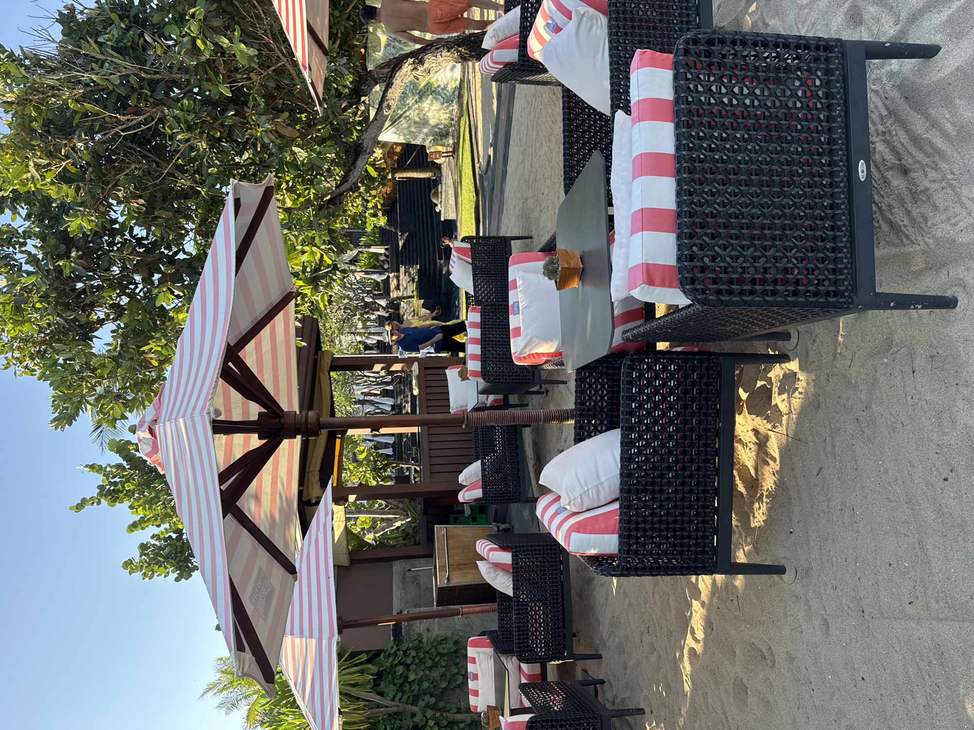 The image shows a beach scene in an unidentified location in Indonesia. The focal point is a set of black rattan chairs with red and white striped cushions and white pillows, arranged near small tables. The chairs appear to be set up as seating for a beachside establishment, possibly a restaurant or resort. Above these seating arrangements are large beach umbrellas with red and white stripes, providing shade. The ground is covered with sand, and in the background, there's a glimpse of a building with a dark roof, possibly a bar or another part of the resort, and some people are visible near it. Lush trees and blue sky are visible in the background.