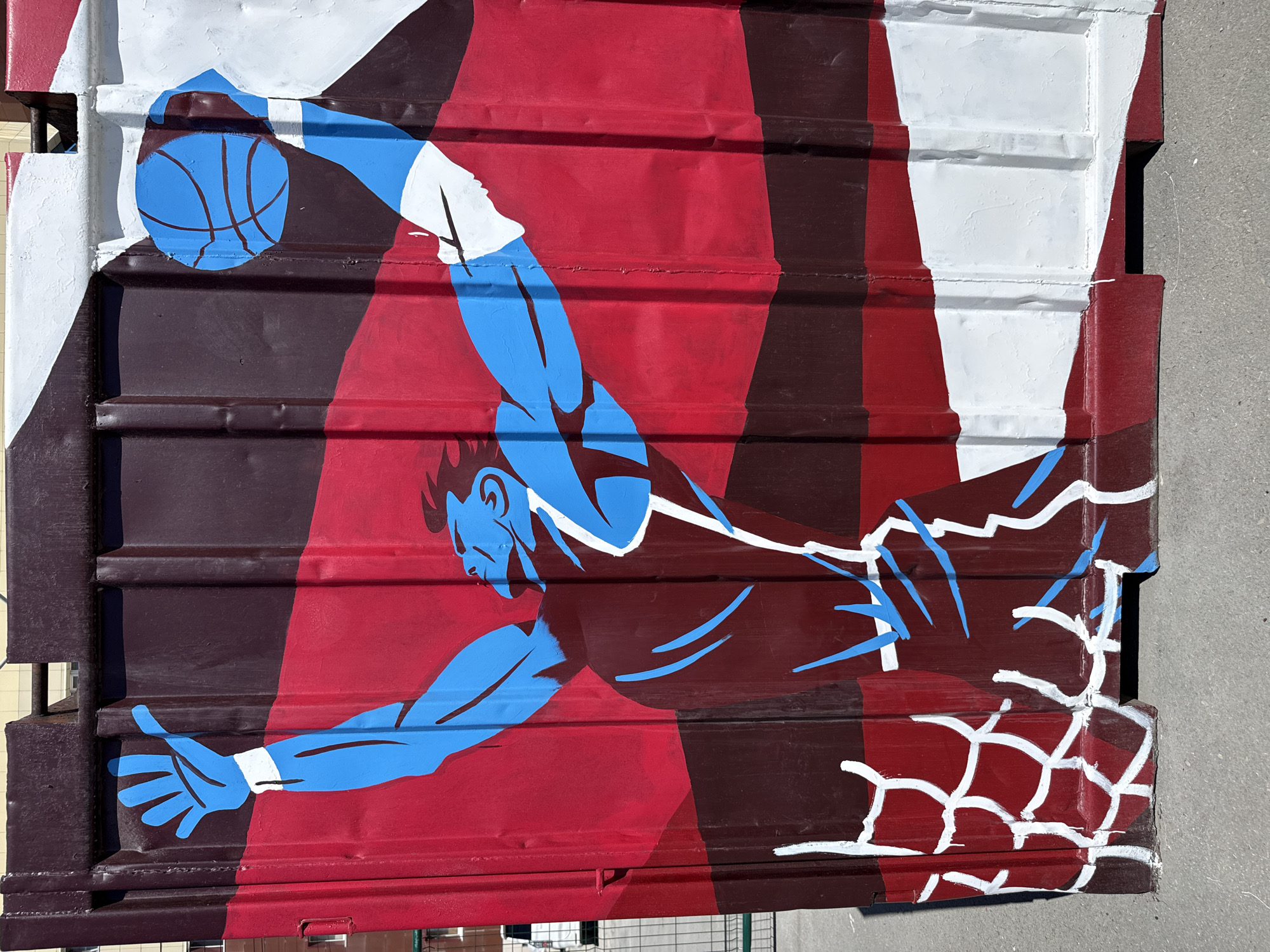 This is a mural painted on corrugated metal, depicting a basketball player in mid-air, dunking a basketball. The player is rendered in a stylized, graphic manner with blue skin and white outlines, wearing a dark red uniform with white accents. The background features bold stripes of dark red, lighter red, and white, creating a dynamic visual. The basketball itself is blue with dark red lines. The net of the hoop is also painted, with white lines on a dark red background.
The mural appears to be an outdoor artwork, likely on a building facade or a similar structure. The lighting suggests it is daytime, possibly with bright sunlight given the shadows and highlights. The corrugated surface of the metal adds texture to the artwork. There is no visible text in the image. The overall mood is energetic and athletic, capturing a moment of intense action. While the specific location within Yakutsk, Russia, isn't identifiable from the image itself, it's a piece of street art in a cold climate, as indicated by the location context.