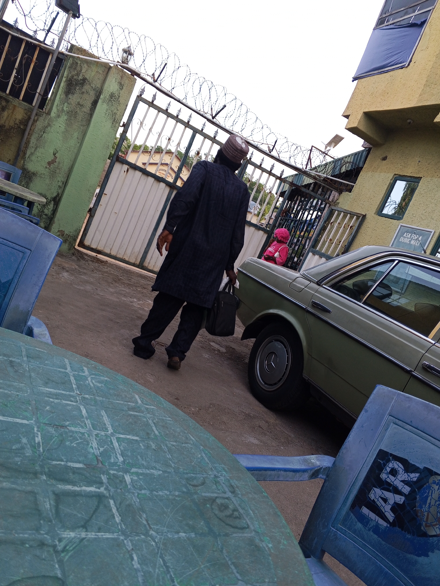 An outdoor scene in Zango-ndaji, Nigeria, captures a man walking away from the viewer on a dusty, unpaved surface. He is seen from behind, wearing a long, dark-patterned tunic or caftan, dark trousers, and a patterned cap. In his left hand, he carries a dark briefcase or bag.
In the midground to the right, a light green/sage-colored vintage car (resembling a Mercedes-Benz sedan) is parked. Further back, near the gate and the car, a small figure, possibly a child, wearing a pink top with a hood and a red backpack, is partially visible.
The foreground features a weathered, teal-colored plastic table and chairs, indicating an outdoor seating area. One chair has a sticker with "TAR" partially visible. A prominent metal gate, topped with barbed wire for security, spans across the upper middle of the frame, leading to an area with another building in the distance, partially revealing the text "APOSTOL... CHURCH".
To the right, a two-story building with a light yellow, textured facade is visible. One of its upper windows is covered with a blue tarp, and a solar panel is mounted on the roof. Below another window on the yellow building, a sign reads "ASK FOR A QUBIC MALI" or "GALL". A security light is attached to a post near the gate, also topped with barbed wire.
The overall lighting suggests daytime, possibly late morning or afternoon, with a bright, overcast sky. The scene depicts an everyday moment in what appears to be a residential or semi-commercial area.