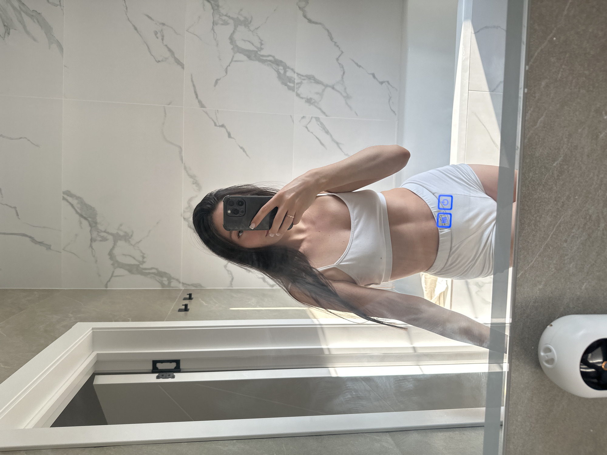 A young woman with long dark hair is standing in a bathroom, taking a mirror selfie with her smartphone. She is wearing a white sports bra and white shorts. The bathroom features marble-patterned tiles on the walls and a white bathtub. A doorway with a white frame and black hinges is visible to the left of the woman. A white object, possibly a security camera or a humidifier, is in the foreground at the bottom of the mirror. There are no visible indications or context within the image to suggest a location in the city of Estachi, Russia.