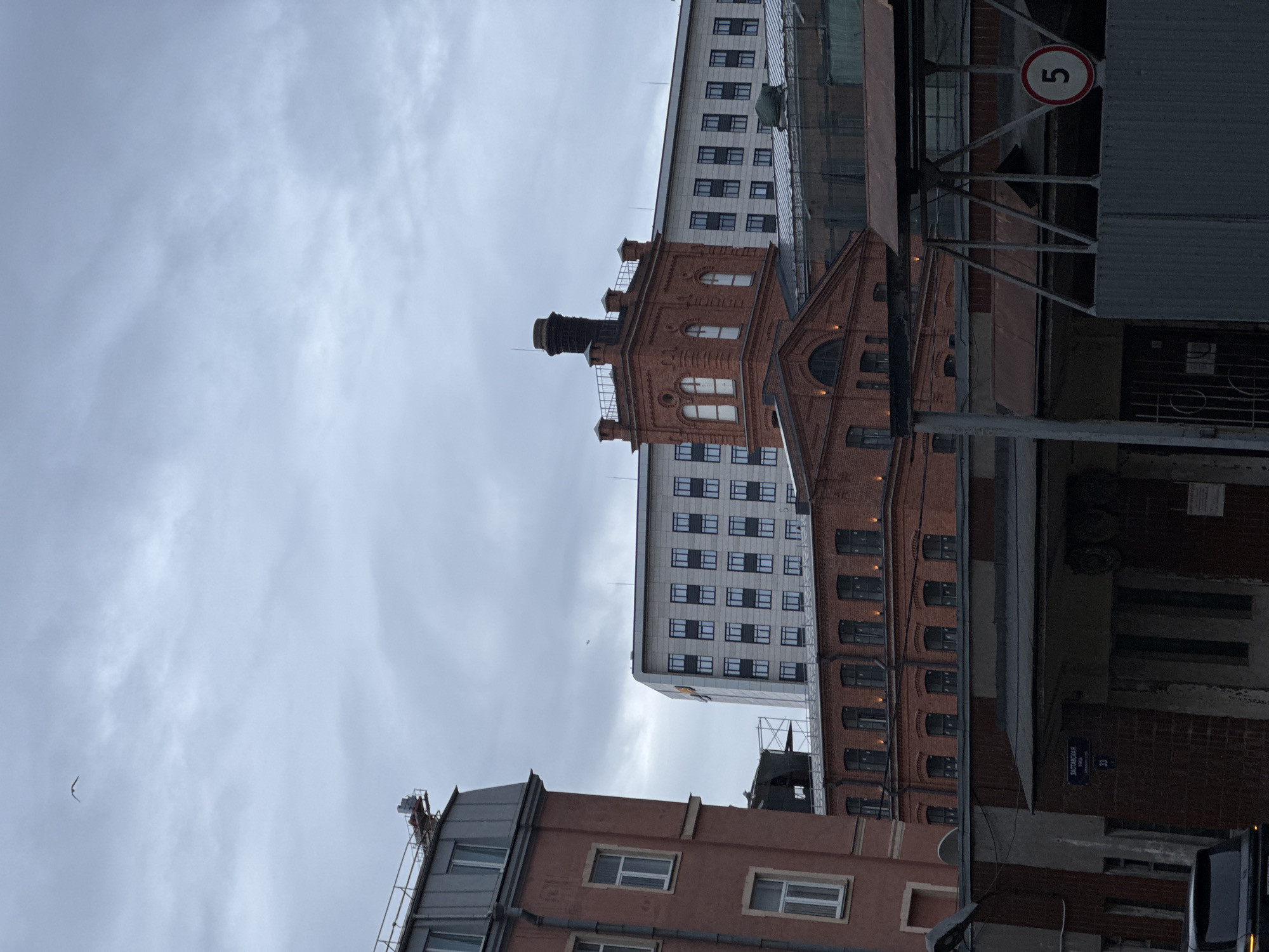The image depicts a street scene in Saint Petersburg, Russia, under an overcast sky. Predominantly, various building facades are visible. An older brick building with distinctive architectural elements like arches and a smokestack stands next to a more modern, taller white building with rows of windows. The juxtaposition of these architectural styles is notable. In the foreground, parts of other buildings are visible, including one with a pinkish hue and smaller windows. A street sign reads "ZASTAVSKAYA ULICA 33," indicating the street name and building number. The presence of a "5" speed limit sign suggests a street or alleyway. Overall, the scene captures an urban environment with a mix of old and new buildings.