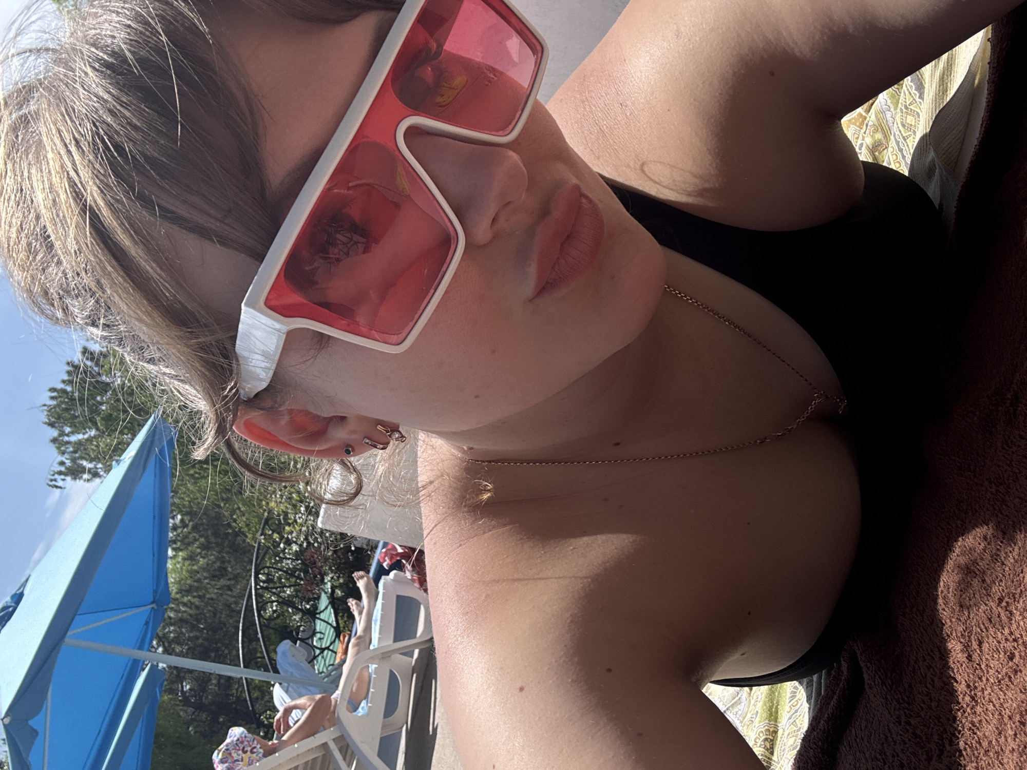 A woman with blonde hair and pink sunglasses is lying on a lounge chair near a pool. She is wearing a black top and a gold necklace. In the background, there are trees, a blue umbrella, and other people lounging by the pool. The location appears to be a resort or hotel pool area. There is no verifiable information to indicate this photo was taken in Yevpatoriya, Ukraine.