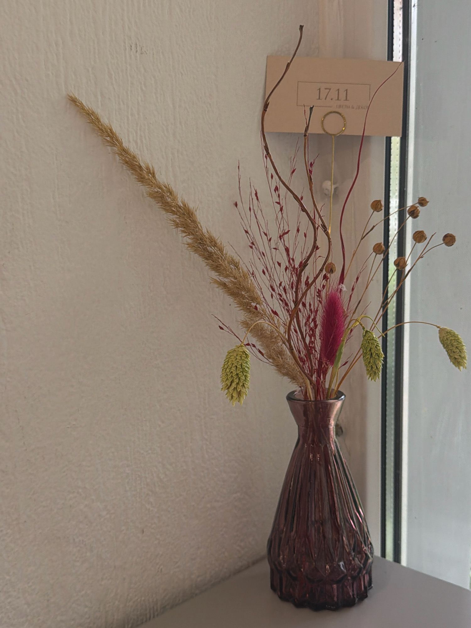 A dark, iridescent glass vase, fluted and with a flared top, holds a dried floral arrangement. The arrangement consists of tall, feathery stalks of beige grass, thin reddish-brown branches with small red buds, a vibrant magenta fluffy stem, and several pale green, cone-shaped seed heads. To the right of the vase, a light brown card is attached to a gold wire easel, displaying the text "17.11" in a simple rectangle at the top. The card also has smaller text beneath, which is illegible in the image. The vase sits on a light gray surface, likely a shelf or tabletop, against a textured, off-white wall. Behind the arrangement and to the right, a vertical window frame with vertical mullions is visible, suggesting an indoor setting with natural light. The scene is devoid of people and action, presenting a still-life composition of decorative elements. The overall impression is one of subdued, naturalistic decoration, possibly in a home or commercial establishment. There is no specific information to ascertain the location within Chaikovsky, Russia, from the visual data provided.
