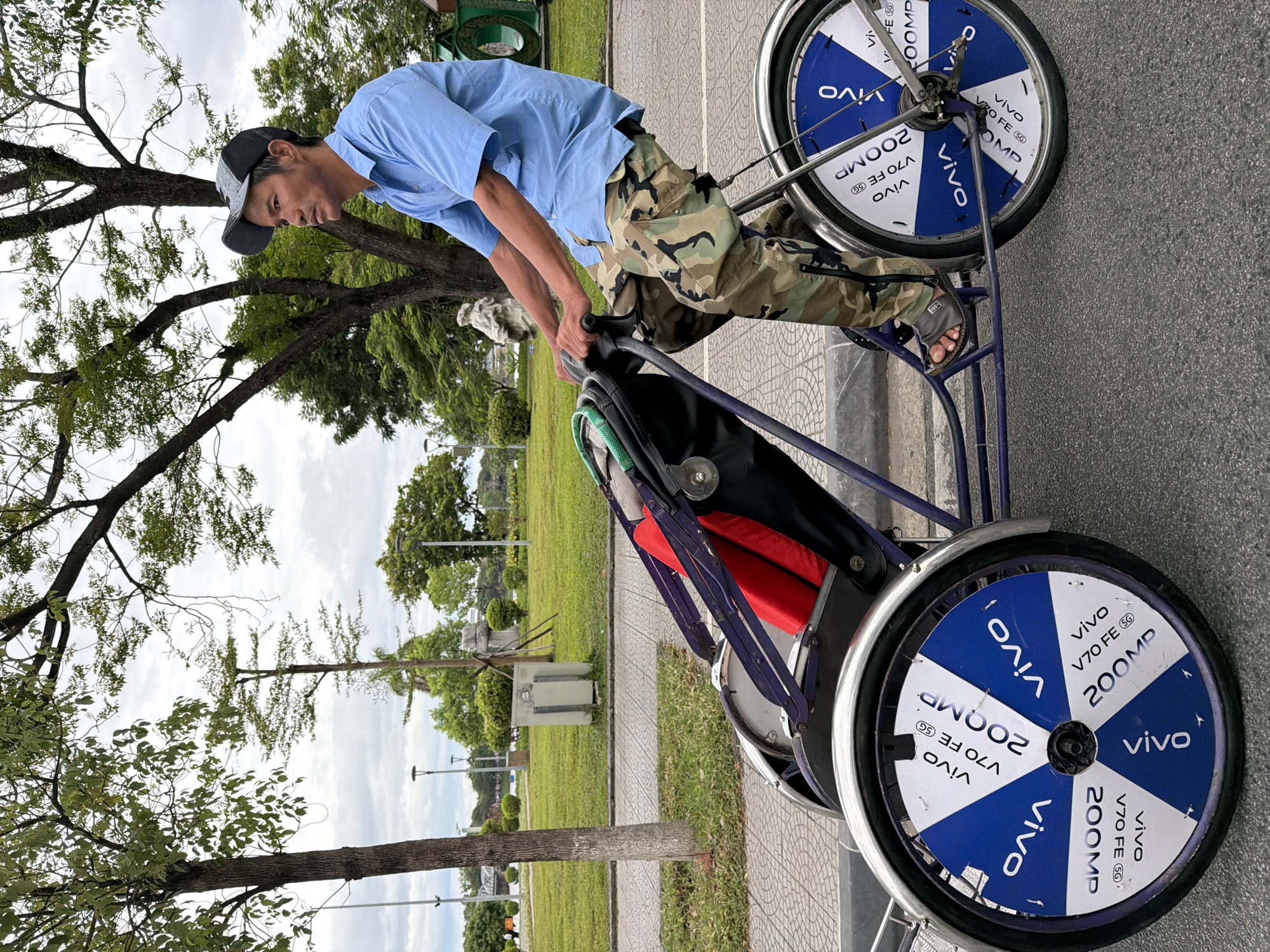 An adult male wearing a light blue short-sleeved collared shirt and camouflage pants rides a tricycle with a passenger compartment. He is wearing a dark baseball cap and sandals. The tricycle has two large wheels on the back, each with a white and blue circular pattern featuring the text "vivo V70 FE 5G 200MP" repeated around the circumference. The passenger compartment is equipped with purple straps and a red cushion. The tricycle is positioned on a paved road next to a sidewalk. In the background, there is a grassy park area with trees, lampposts, and a body of water. The sky is overcast. The image appears to be taken in Thuận Hóa Ward, Vietnam, likely in a park or along a waterfront promenade.