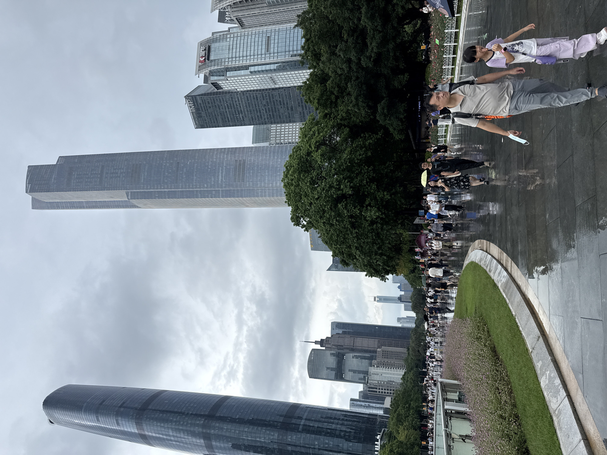 In Tianhe District, China, on a cloudy, wet day, a paved walkway curves through a park-like setting. The wet pavement reflects the overcast sky and the surrounding environment. A large crowd of people walk along the pathway, some carrying umbrellas, suggesting recent or ongoing rain.
Dominating the background are several modern skyscrapers. To the left, a tall, curved skyscraper with dark, reflective glass rises into the sky. To its right, a much taller, rectangular skyscraper with a similar glass facade stands prominently. Other buildings of varying heights and architectural styles are visible between and beyond these two structures, forming a dense urban skyline.
In the foreground, a man and a young girl walk together on the wet path. The man, wearing a light-colored polo shirt and gray pants, carries a light blue face mask in his hand and a backpack. The girl, dressed in a purple t-shirt and light purple pants, is sticking out her tongue and holding a lollipop. They are walking away from the viewer and towards the right side of the frame.
To the left of the walkway, a patch of green grass is bordered by a stone curb. Beyond the grass, a bed of small, pink flowers lines a decorative metal fence. Further to the left, a curved, grey structure with a metal railing appears to be part of a pedestrian barrier or enclosure. Lush green trees are scattered throughout the scene, interspersed with the buildings and the walkway, adding a touch of nature to the urban landscape. The overall impression is of a busy public space on an overcast day in a modern Chinese city.