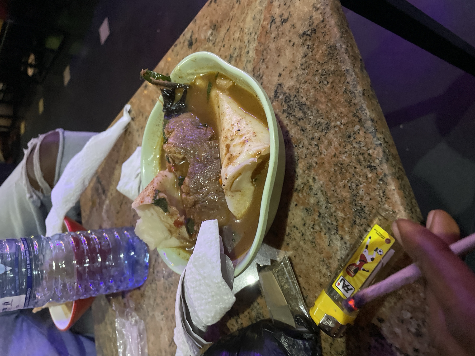 The image depicts a scene on a marble-patterned table. A bowl of soup with meat and what appears to be a wrap or dumpling sits in the center. To the left of the bowl is a bottle of water and a crumpled tissue. To the right of the bowl, a hand is holding a lit cigarette, with a yellow lighter beside it. A partially visible object that is black is under the tissues. Based on the details visible in the bottle, this photo was taken in Obosi, Nigeria.