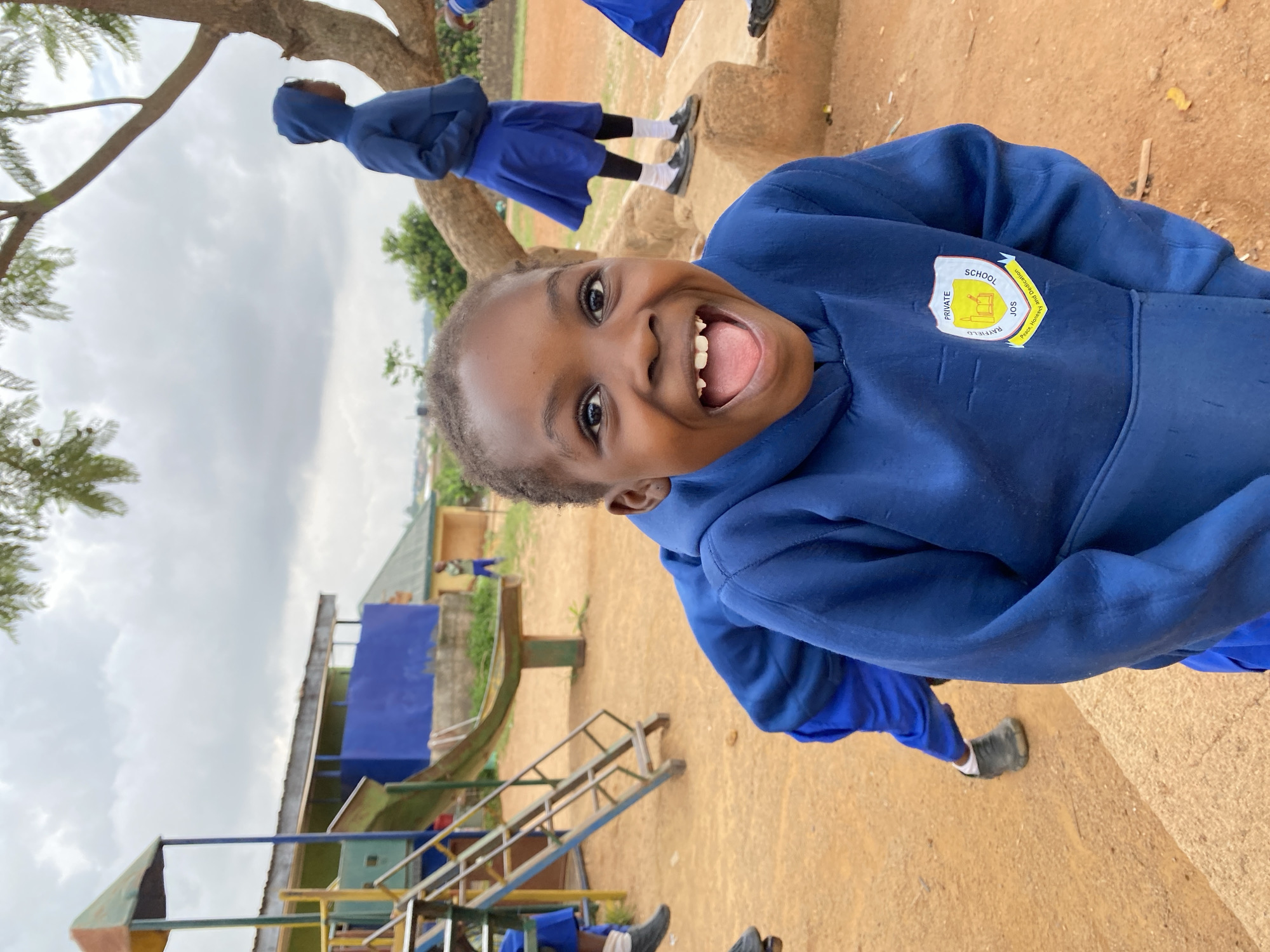 A young, dark-skinned child with short hair, wearing a blue hooded sweatshirt with a school patch on the chest, is smiling widely with an open mouth, showing teeth. The patch reads "Rayfield Private School Jos". The child is outdoors on a playground in Gura Top, Nigeria. The playground includes a metal slide and climbing structure. Other children in similar blue school uniforms are visible in the background, along with trees and a building. The sky is overcast. The ground appears to be reddish dirt.