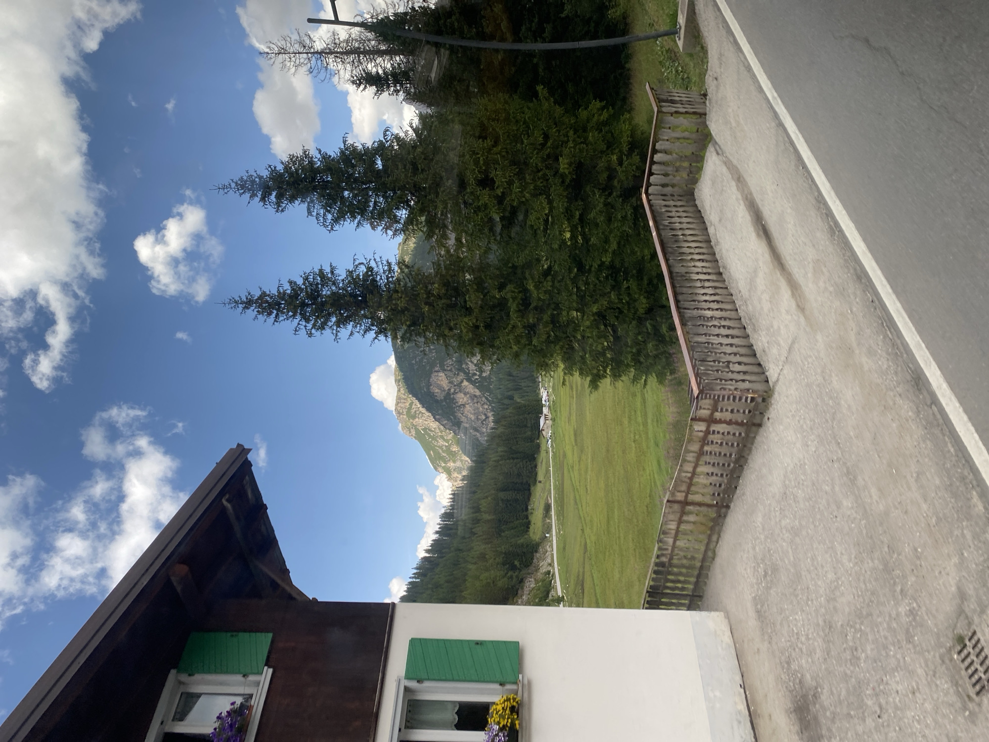 The image depicts a scenic view of the Dolomites in Auronzo di Cadore, Italy. In the foreground, a white building with dark wooden accents and green shutters on its windows is partially visible on the left. The building has a rustic aesthetic. A concrete path leads away from the building, bordered by a weathered wooden fence. Beyond the fence, a lush green meadow stretches out, dotted with a few trees and a road that curves through the landscape. In the mid-ground, a dense forest of evergreen trees covers the foothills of the mountains. The background is dominated by majestic, rugged mountains under a bright blue sky with scattered white clouds. A few small buildings and parked cars can be seen in the distance in the valley. The overall scene conveys a sense of tranquility and natural beauty characteristic of the alpine region.
