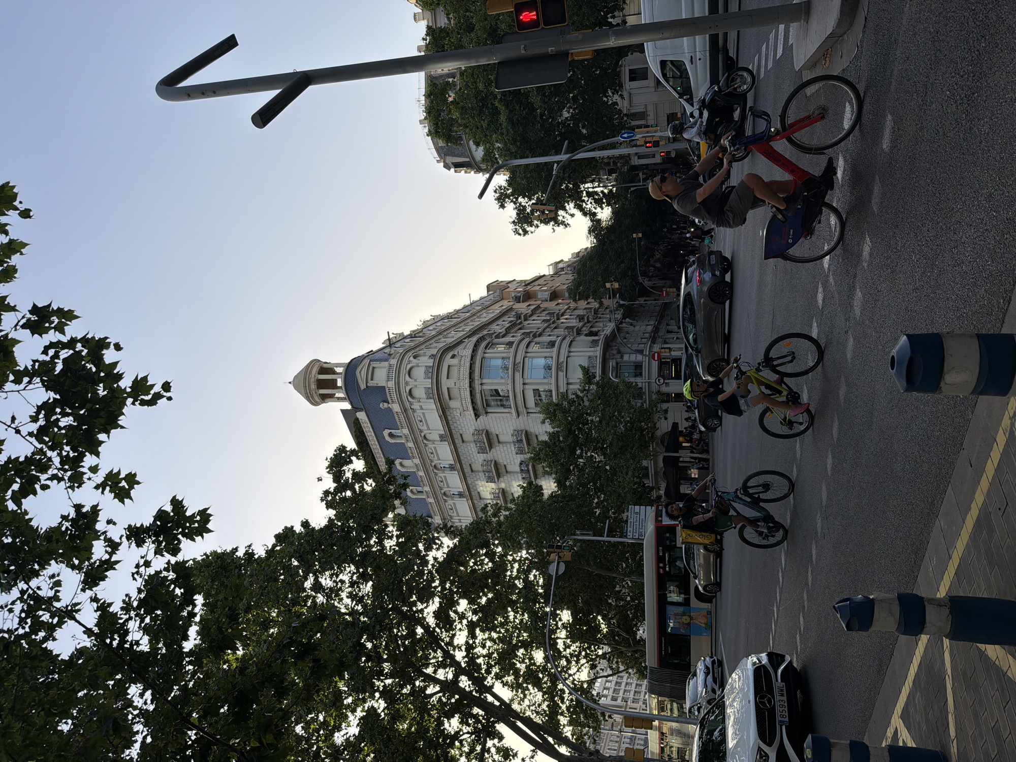 The image captures a street scene in Barcelona, Spain, with a prominent building in the background and several cyclists in the foreground. The building is large and ornate, with a rounded corner and a distinctive domed roof feature, suggesting an older, grand architectural style common in European cities. Trees with dense foliage flank the building and extend into the upper left quadrant of the image, their leaves creating a dappled effect with the sunlight.
In the foreground, the asphalt street is marked with white lines indicating traffic lanes and a pedestrian crossing. Several individuals are seen riding bicycles. One person, wearing a yellow helmet, is on a light-colored bicycle. Another cyclist, wearing a green helmet, is on a darker bicycle. A third cyclist, wearing a baseball cap and sunglasses, is on a red and blue bicycle. The cyclists appear to be moving across the intersection.
A white Mercedes car with license plate "8593 MML" is visible on the left side of the road, having just crossed the white lines. A bus, partially obscured by trees, is also present in the background. A white van and a motorcycle can be seen further down the street. Traffic lights are visible, with one showing a red pedestrian signal. The sky is clear and pale, indicating either early morning or late afternoon light. The overall impression is a busy urban environment with a focus on sustainable transportation, as evidenced by the number of cyclists.