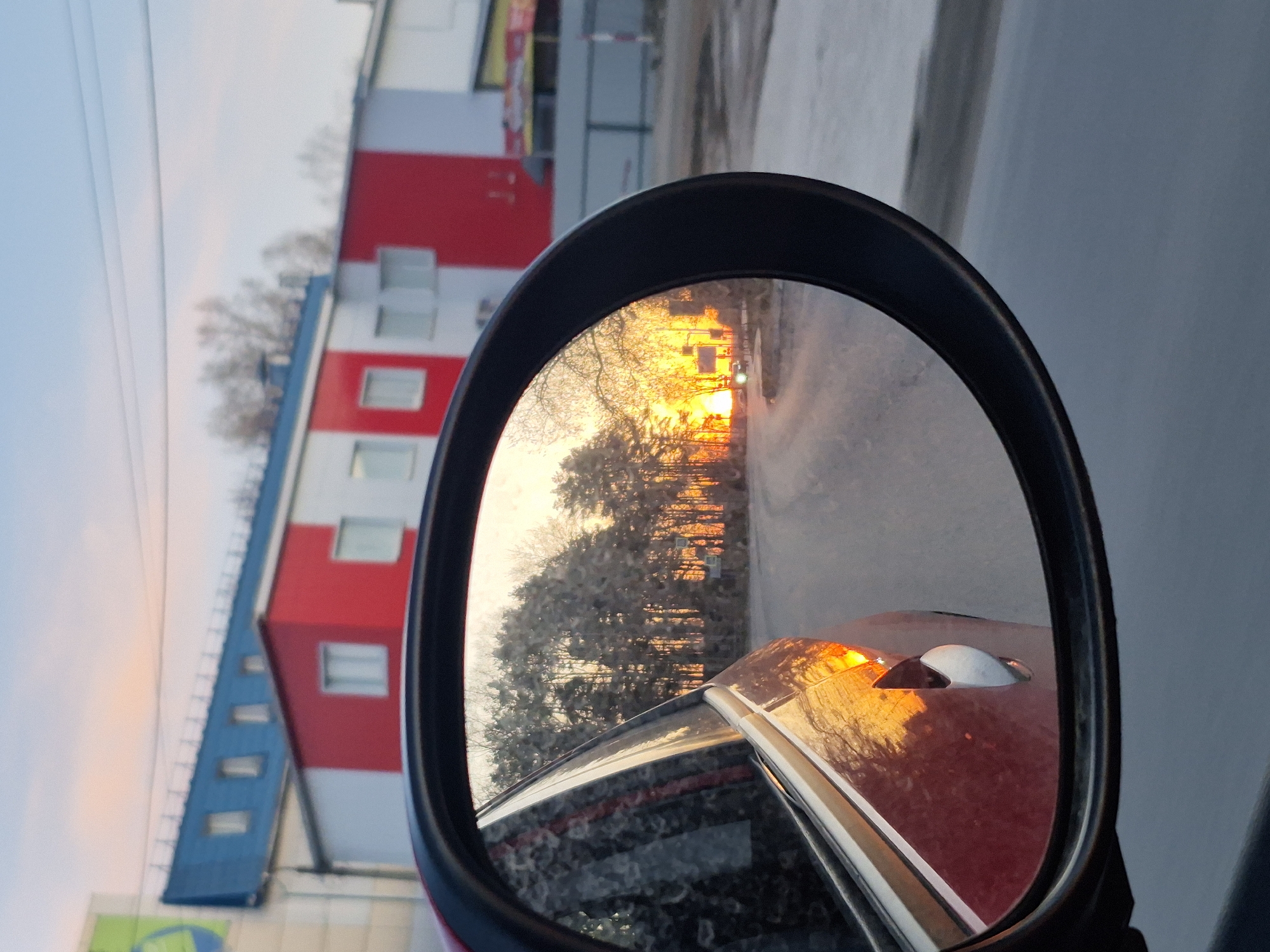 The image depicts a side-view mirror of a car, reflecting a sunset and a road with trees. The car itself is red. In the background, outside the mirror's reflection, is a building with red and white stripes, as well as a blue roofline. The image appears to be taken at dusk or dawn. The surrounding environment suggests an urban or suburban setting. No people are visible. The location is in the city of Chaikovsky, Russia.