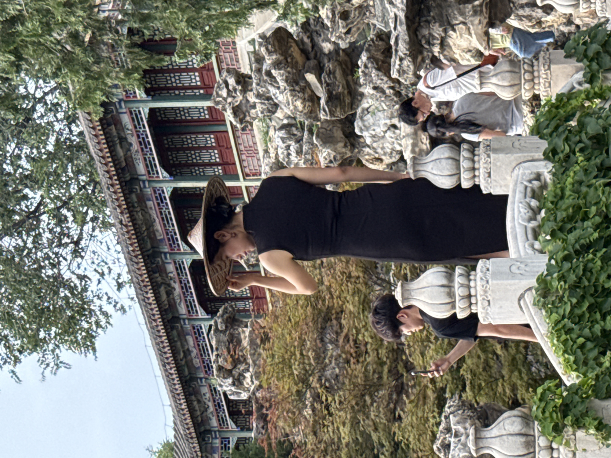 In the Xicheng District, China, at what appears to be a traditional Chinese garden or park, a woman in a black dress and a conical straw hat stands on a white stone balustrade, adjusting her hat. She is the primary subject in the foreground. To her left, a young man in a black t-shirt is captured in profile, holding a smartphone and appearing to take a photo. Behind and to the right of the woman, a man in a white shirt with glasses and a dark bag slung over his shoulder is visible, along with a woman in a grey t-shirt. Further to the right, another person in a green shirt and blue pants is partially visible. The background features a multi-tiered building with ornate red and blue decorative elements and a tiled roof, as well as rocky landscaping and lush green trees. The overall scene is one of people visiting a cultural landmark.