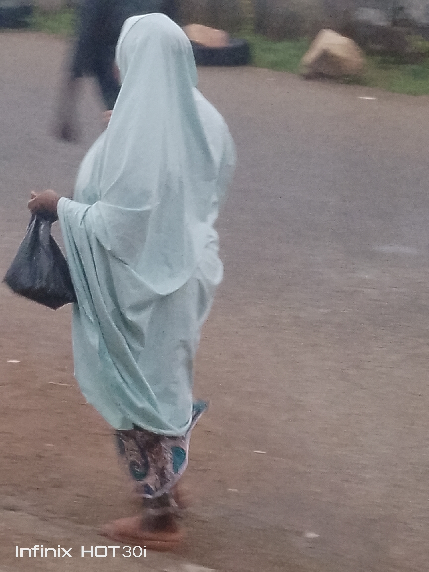 A woman wearing a light blue hijab and traditional clothing is captured from the side, walking on an asphalt surface. She is holding a black plastic bag in her left hand. Her patterned lower garment is visible below the flowing fabric of her outer clothing. In the background, out of focus, a person in dark clothing is walking away. The environment appears to be outdoors with some greenery and rocks visible in the distance. The bottom left corner of the image displays "Infinix HOT 30i".