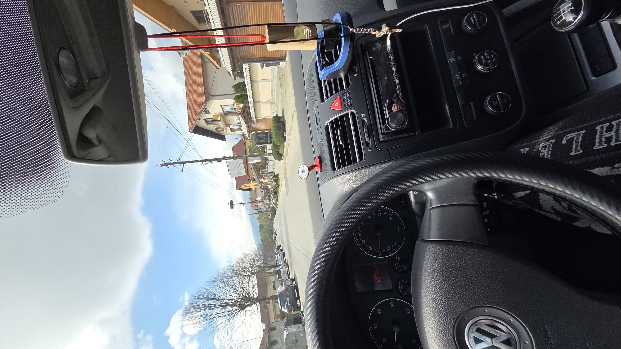 The image captures a first-person perspective from inside a car, looking out onto a residential street. The sky is bright blue with white clouds, indicating daytime.
Inside the car, the dashboard and steering wheel dominate the foreground. The steering wheel features a textured, carbon fiber-like wrap on its rim and a prominent Volkswagen logo on the horn pad. The dashboard includes air vents, a red triangular hazard light button, and an aftermarket radio/media player. Below the radio, buttons labeled "AUTO," "DUAL," and "ON/OFF" suggest climate control. A manual gear stick with a "1-6" pattern is visible to the right. A blue phone holder, a small wooden block, and a key are suspended near the rearview mirror. A small, red circular mirror is attached to the dashboard. The instrument cluster shows a speedometer and tachometer with readings like "20," "40," "60," "220," "240," and "260 km/h." An odometer displays "50000" (likely 50,000 km), and a digital display shows "15:14." Other visible text includes "PASSENGER AIR BAG OFF" and "ESP" indicators. Part of a patterned blanket or cover with "T H L" text is seen on the passenger seat.
Outside the car, a paved road extends into the distance, lined with parked vehicles on the right. Various houses with pitched roofs and different architectural styles populate both sides of the street. Utility poles with numerous power lines run along the road, one of which prominently features a large stork nest. A large tree with bare branches stands on the left, possibly indicating late winter or early spring. Hills or mountains are visible in the distant background, completing the serene, everyday scene of a town or village.