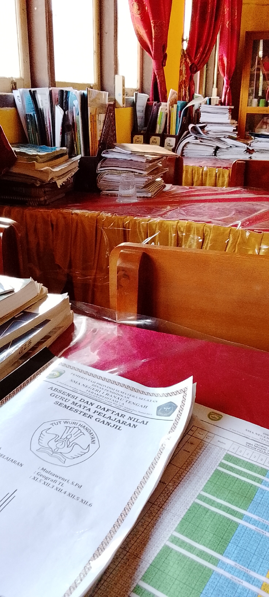 This is a close-up, slightly angled, eye-level shot of a desk or table surface filled with educational materials and furniture. The dominant colors are the warm tones of wood, the deep red of the table cloth, and the white and yellow of papers and curtains. The scene appears to be indoors, likely in an educational institution, given the presence of books, binders, and documents with school-related headings.
In the foreground, a white document with "ABSENSI DAN DAFTAR NILAI SEMESTER GANJIL" (Attendance and Grade List, Odd Semester) is prominently displayed, along with the "TUT WURI HANDAYANI" logo, a symbol of Indonesian education. Below this, there are handwritten notes indicating a teacher's name ("Mulzawanri, S.Pd") and subjects ("Geografi TL", "XI.5, XII.3, XII.4, XII.5, XII.6"). To the right of this document, a portion of a timetable or grading sheet is visible, featuring a grid of green and white cells with handwritten entries.
The middle ground reveals stacks of books and papers on a red tablecloth that covers the table. A clear glass with some liquid sits atop one stack of papers. Further back, a wooden desk or partition is visible, with more stacks of documents and books arranged on it. The background features a window with wooden frames and red curtains, suggesting natural light is entering the room. The overall impression is one of an organized, yet busy, workspace.
The image does not provide specific details or visual cues that would allow for the identification of the city of Jepara, Indonesia.