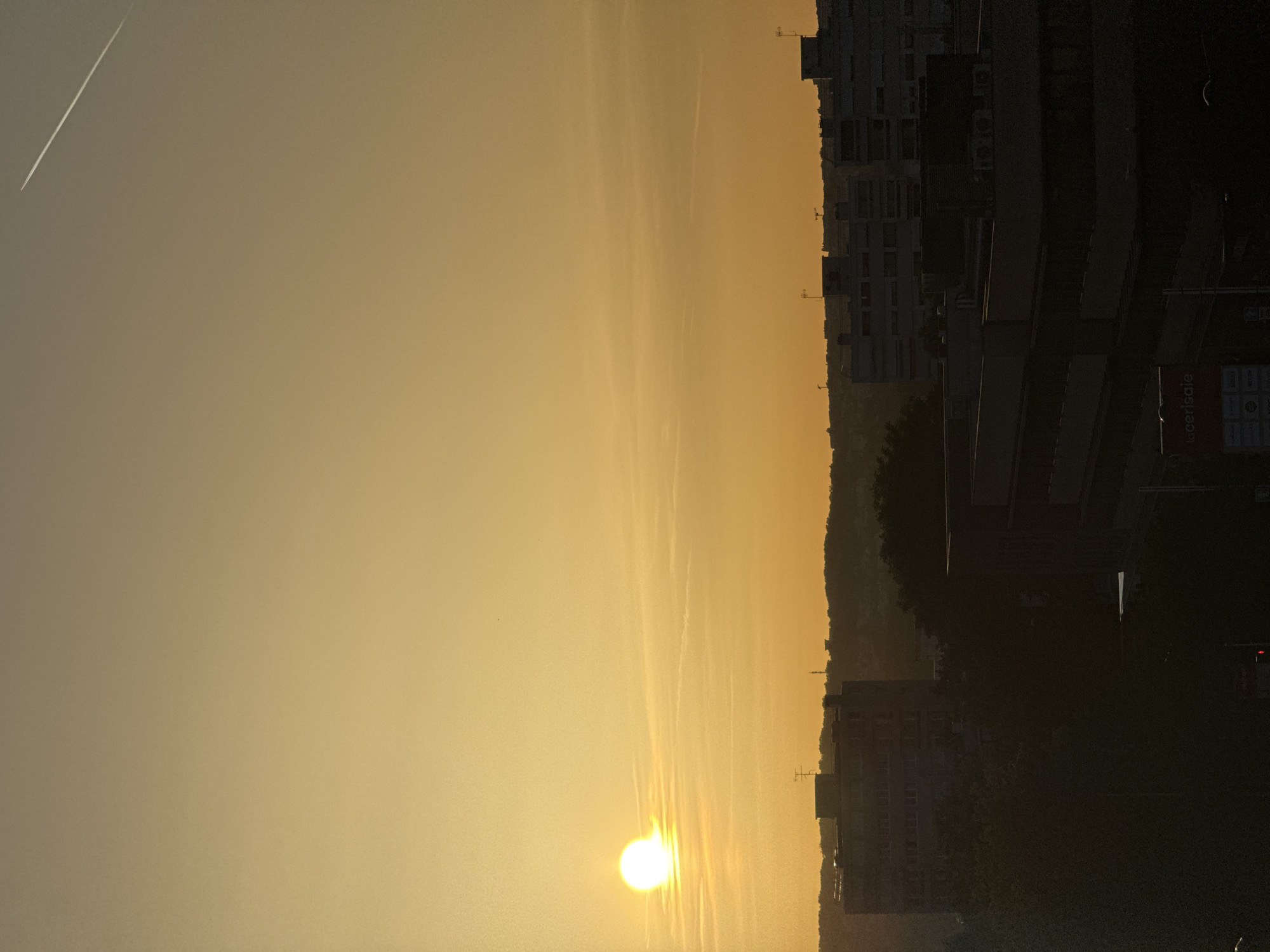 An outdoor, vertical, eye-level medium shot captures a sunset over a city. The sun is a bright, white orb on the left side of the frame, partially obscured by thin, wispy clouds that stretch across the sky. The sky transitions from a pale yellow near the horizon to a lighter yellow at the top. A faint contrail from an airplane is visible in the upper right corner.
The foreground is dominated by the dark silhouettes of buildings and trees. A tall, multi-story building with visible window rows stands prominently in the mid-left, topped with an antenna. To its right, a series of lower-rise buildings with flat roofs and what appear to be rooftop antennas are silhouetted against the horizon. More buildings, dark and indistinct, fill the lower portion of the frame, with a sign that reads "laCerisaie" visible on one of them. The overall scene depicts an urban landscape bathed in the warm, fading light of sunset.