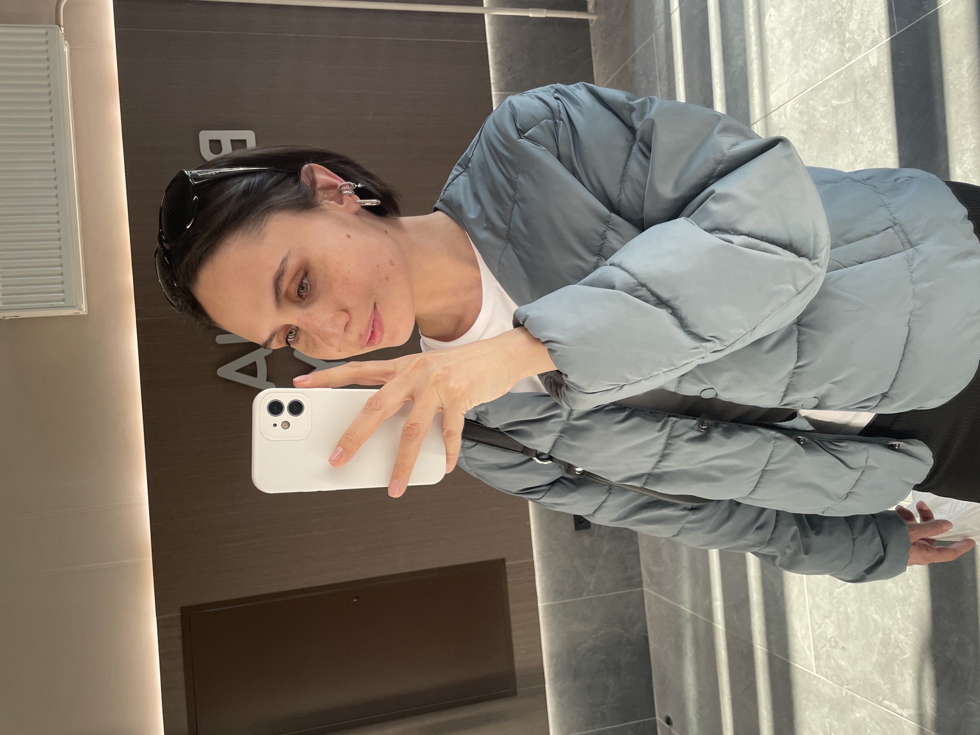 A woman is taking a selfie in what appears to be a building in Pervomaysky, Russia. She is wearing a light blue puffer jacket over a white t-shirt. Sunglasses are perched on her head. Her phone is white and held in her right hand. The background includes a brown wall with some signage and a reflective surface showing the vertical lines of a tiled wall.