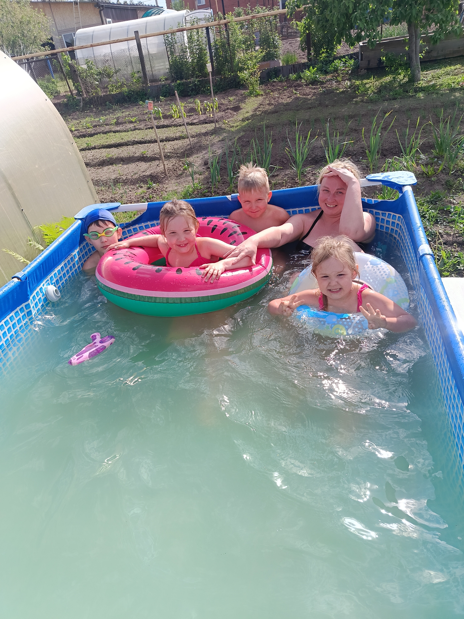 A high-angle, full shot captures a rectangular above-ground swimming pool with a blue frame and patterned blue and white interior lining, located in a garden or backyard in Chaikovsky, Russia. Five individuals are in the pool. On the left, a boy wearing a blue swimming cap and green-tinted goggles is partially submerged. Next to him, a girl in a pink bathing suit floats in a pink and green watermelon-shaped inner tube. To her right, a boy with blond hair looks towards the camera. Behind the children, a woman in a black bathing suit is partially submerged with her hand on her head. In the front right corner, a girl in a pink bathing suit floats in a blue inner tube and is making a peace sign with her hands. A pink and purple toy rests in the water near the first child.
Behind the pool is a garden with rows of plants, including what appears to be garlic, and a small greenhouse to the left. In the background, a fence separates the property from neighboring buildings. The sky is partially visible, suggesting a sunny day.