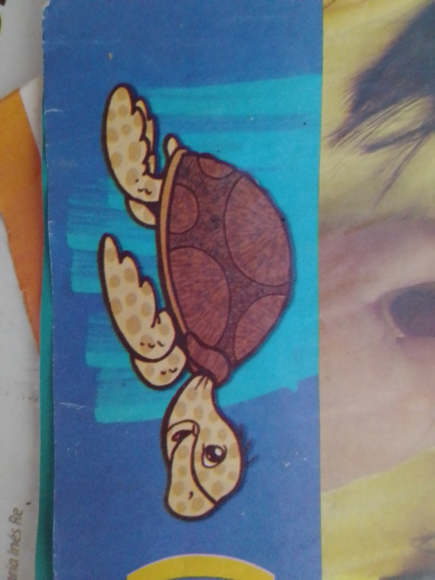 The image shows a close-up, vertical, slightly angled shot of a cartoon drawing of a turtle on a blue background. The turtle is drawn in a simple, whimsical style with a brown shell, yellow flippers, and a yellow head with a black eye and mouth. The turtle appears to be swimming as its flippers are outstretched. The drawing is on what appears to be a card or sticker. To the right of the drawing, a portion of a person's face is visible, appearing out of focus and in a warm tone, possibly from a photograph or another printed image. There is also some text on the left side of the image, partially obscured, which appears to read "aria Inés Re". The image does not provide any information about the location in the city of La Tagua, Venezuela.