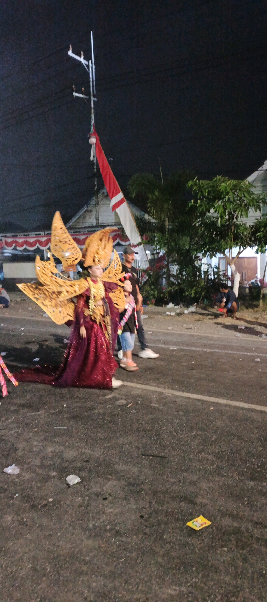 In Kasiyan, Indonesia, a parade takes place at night. A person in a vibrant, ornate costume, adorned with large golden wings and a rich red gown, stands prominently on the road. Beside them, two individuals, possibly performers or escorts, are also present. One is an adult male wearing a dark shirt and a baseball cap, while the other is a child in a black t-shirt and denim shorts, also decorated with a sash. The Indonesian flag, red and white, flies high on a utility pole in the background, partially obscured by the night sky and electrical wires. A few trees are visible to the right, with buildings and possibly tents lining the street. The road itself is paved and shows signs of wear, with scattered debris like plastic wrappers. In the distance, another person is crouched down on the side of the road. The atmosphere appears festive, given the parade context and the decorative costumes.
