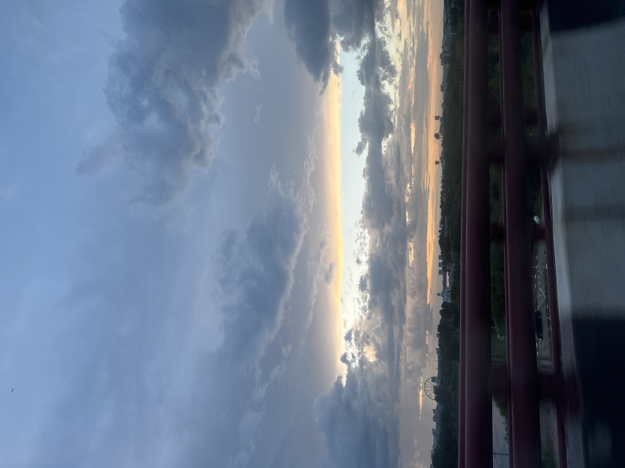 The image captures a dramatic sky over a city at dusk, likely Irkutsk, Russia. A band of warm light, transitioning from yellow to pale blue, cuts across a dramatic expanse of dark, heavy clouds. The clouds are textured and varied, with some appearing wispy and others dense and ominous.
Below the sky, a silhouetted cityscape stretches across the horizon. A Ferris wheel is prominently visible on the left, a common landmark in many cities. Trees form a dark band between the cityscape and the foreground. The foreground is dominated by a dark red railing, suggesting the photo was taken from a bridge or elevated walkway. The railing is blurred, indicating motion, possibly from a vehicle. The surface below the railing is reflective, possibly wet from recent rain. The overall mood is one of atmospheric transition, with the vibrant colors of the sunset struggling to break through the brooding clouds.