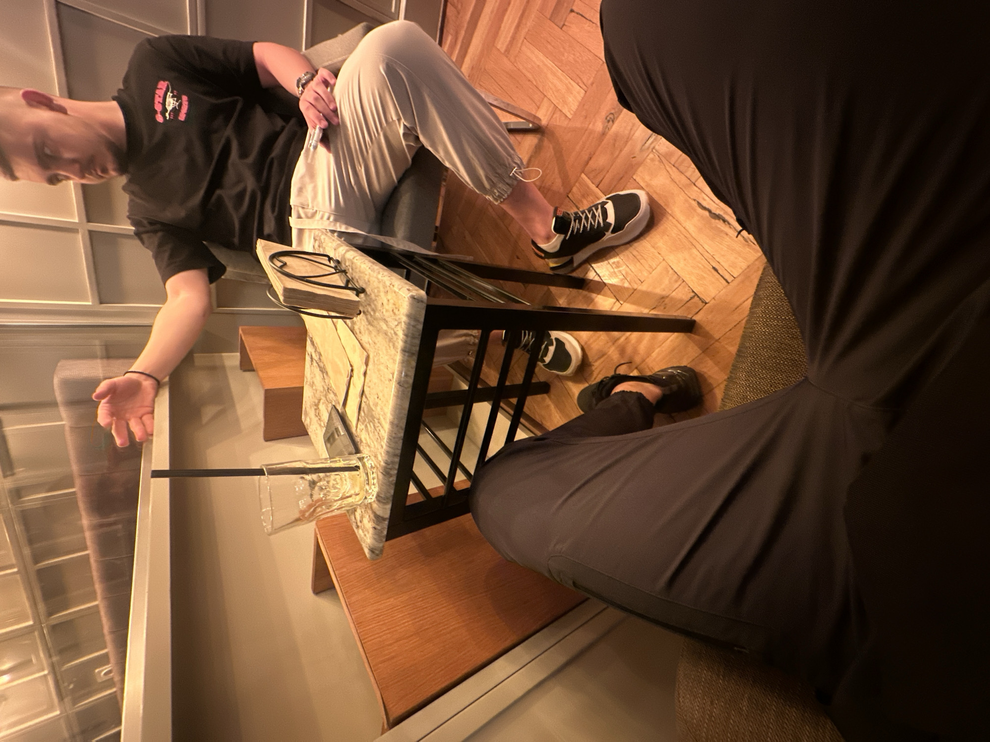 The scene appears to be indoors, perhaps a bar or lounge. A person wearing a black t-shirt and light-colored pants sits at a small table. Another person, mostly obscured, is wearing black clothing. A glass with a straw is on the table. The floor is wooden in one area and there are steps and a window in the background. Without additional information, pinpointing the exact location in Burgas, Bulgaria is impossible.