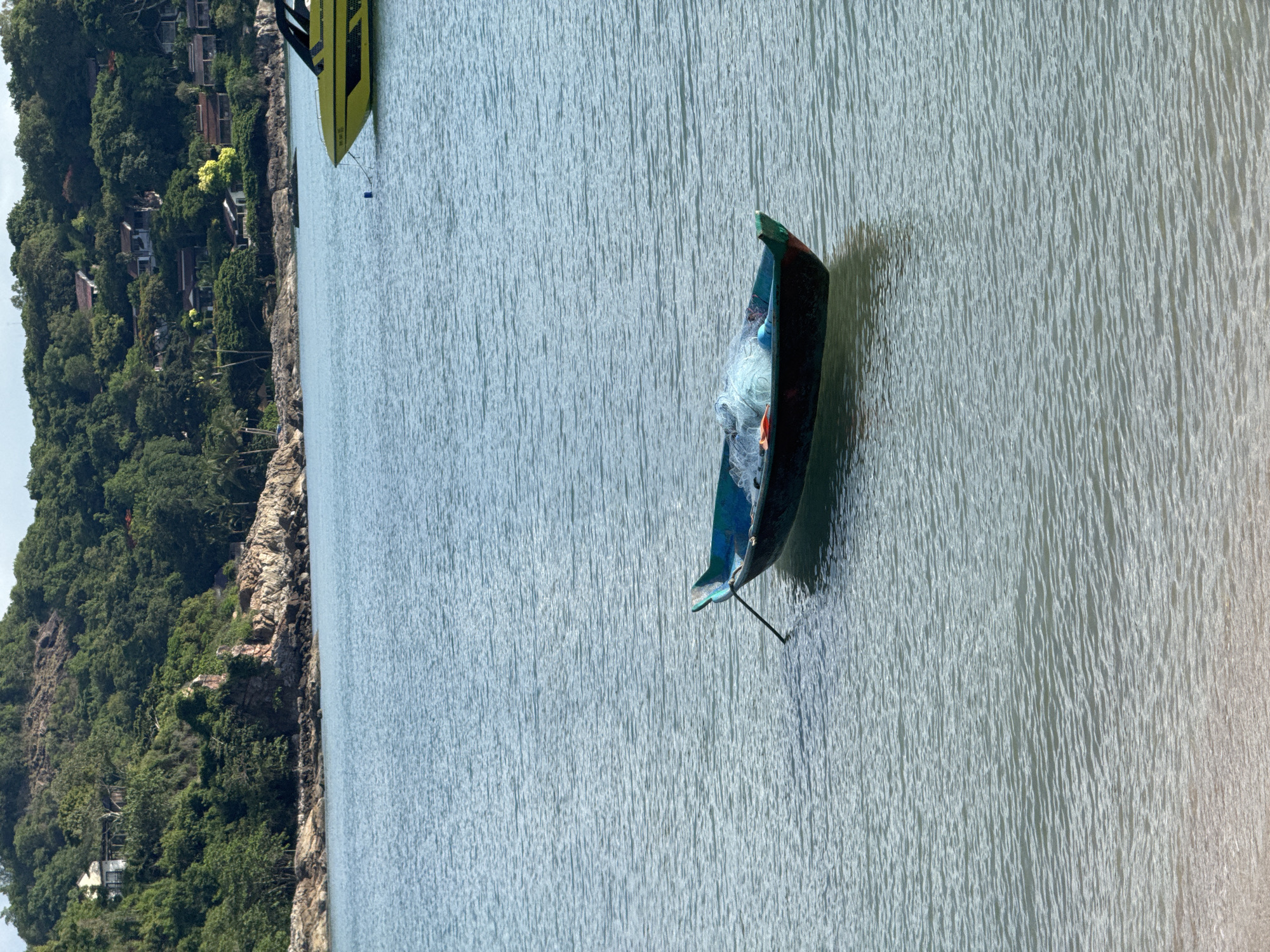 In the image, a small, dark-hulled boat with a green trim rests on calm, blue water. A fishing net is piled inside the boat. The boat is tethered to something unseen by a rope and oar. In the background, a yellow speedboat is moored in the water. Beyond the water, a densely forested hillside with several houses nestled among the trees rises up. The sky is a light blue with some white clouds. The foreground shows a sandy beach leading into the water.