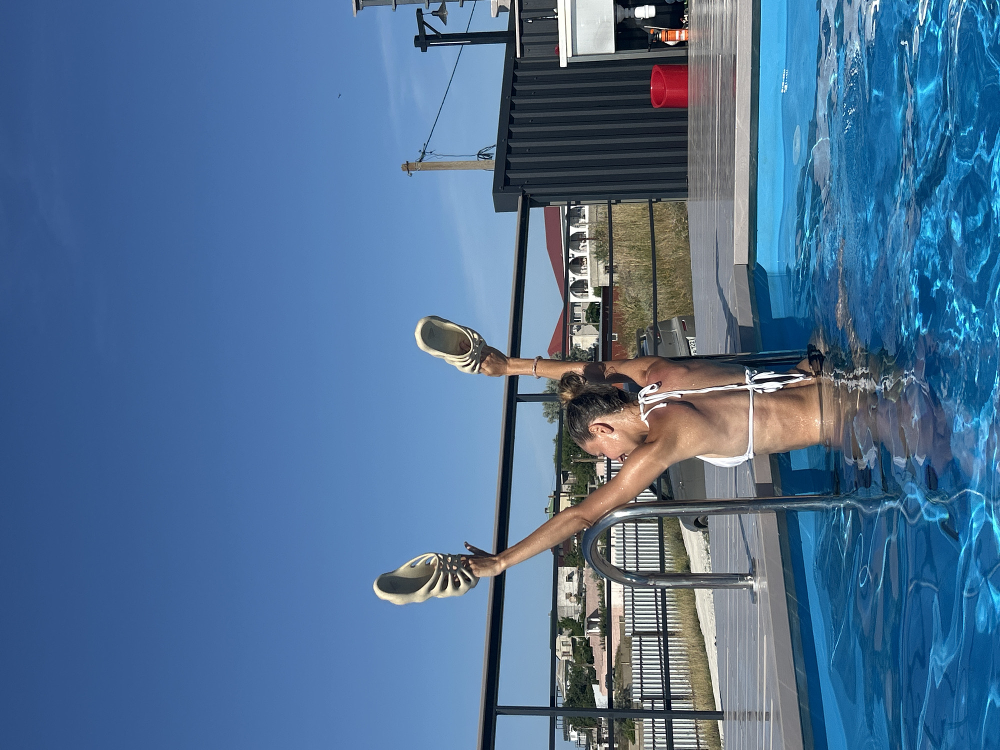 A woman in a white bikini is standing in a bright blue swimming pool, holding a pair of cream-colored slides above her head. She is positioned near the pool ladder, with her back to the camera and her arms raised. The water is clear and reflects sunlight. The pool is surrounded by a wooden deck and a metal railing. In the background, there are buildings with red roofs, a fence, and a clear blue sky. The scene appears to be outdoors, likely in a residential area. The location is identified as Mysivske settlement, Ukraine.