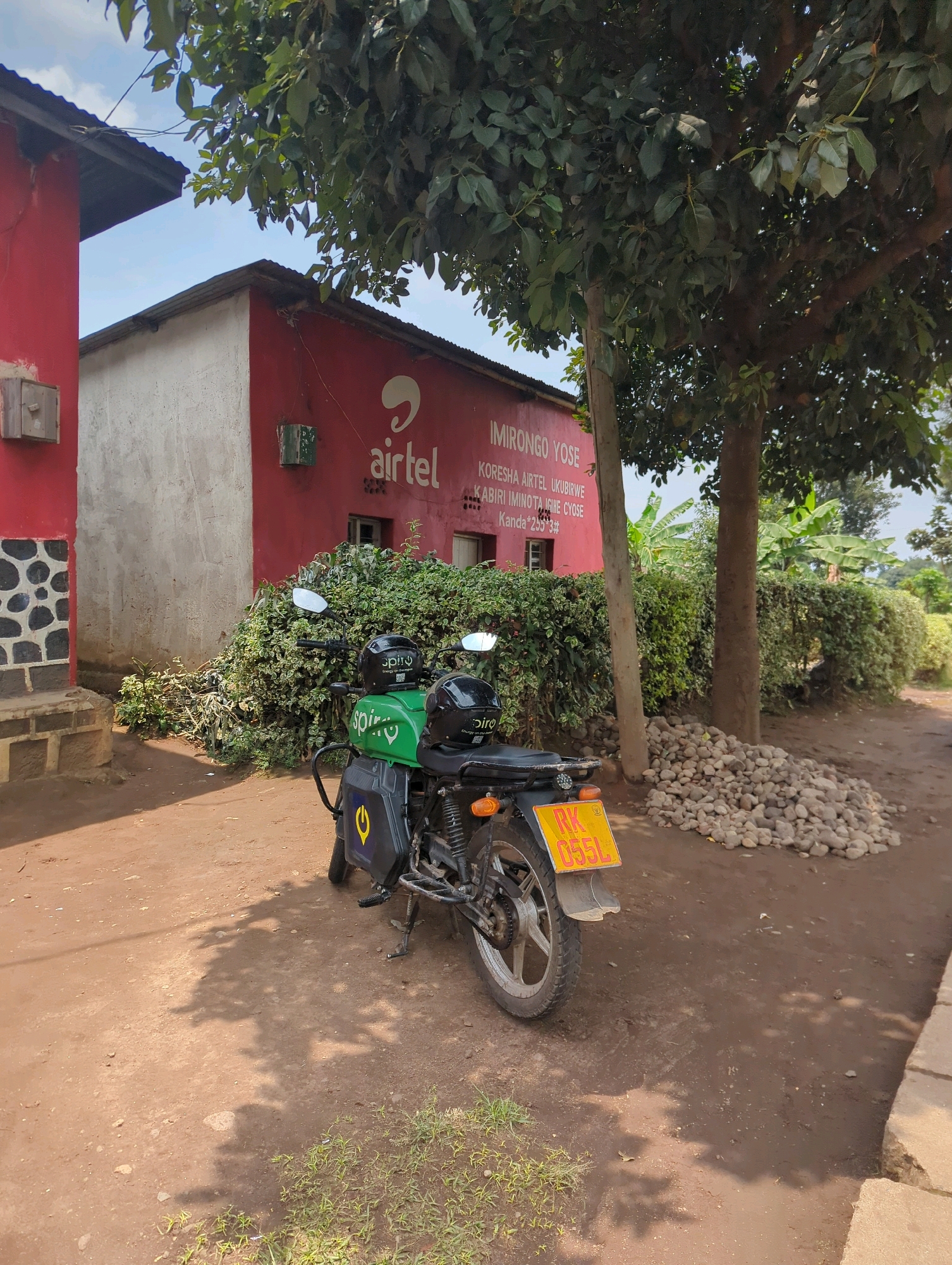 A red and white building with a corrugated metal roof has an Airtel logo and text in Swahili on its red facade. In front of the building, a motorcycle with two helmets and a green cargo box is parked on a dirt road. A pile of rocks is located to the right of the motorcycle, and a line of green bushes runs along the building. The image appears to be taken in Musanze District, Rwanda.
