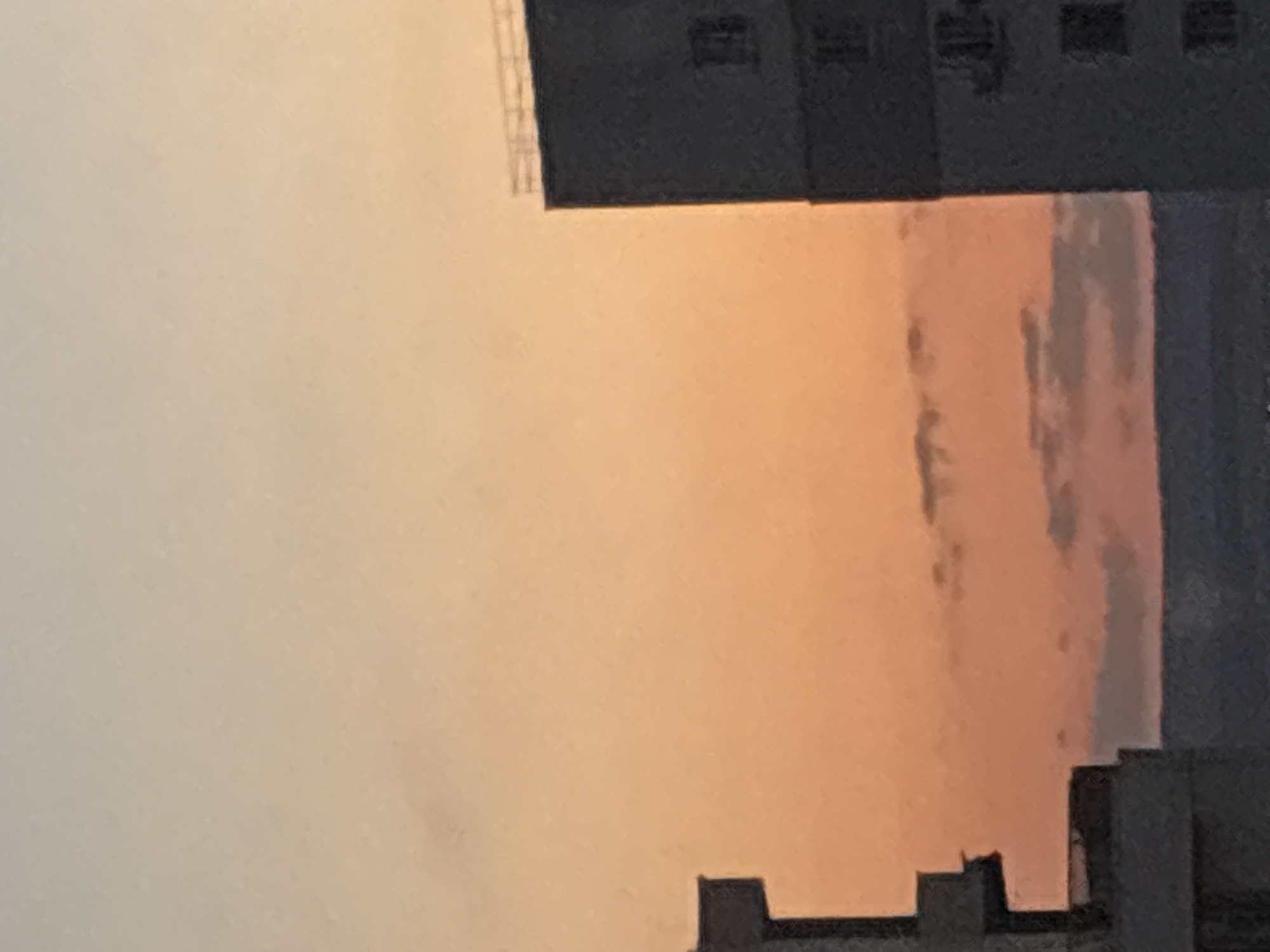 The image depicts a cityscape at dusk with a gradient sky of pale orange and yellow hues. The silhouettes of two multi-story buildings are visible against the sky. The building on the right is taller and darker, with rectangular window-like structures visible on its facade. The building on the left is partially obscured and appears to have a more complex architectural shape with protruding sections. A faint horizontal line, likely the horizon or another distant structure, is visible at the bottom. The scene suggests an urban environment in Sofia, Bulgaria, characterized by residential or office buildings against a twilight sky.
