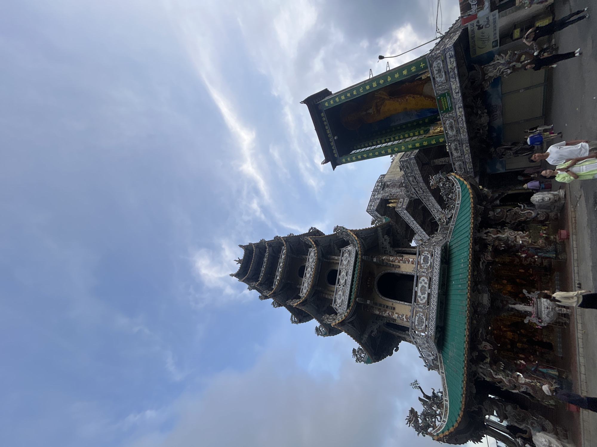 The image, captured from a low angle and rotated sideways, features a highly ornate Buddhist temple complex in Xuân Thành, Vietnam, under an overcast sky.
The central focus is a tall, multi-tiered pagoda, intricately detailed with numerous sculptures and decorative elements. Each level of the pagoda is topped with a green-tiled roof, contributing to its vibrant appearance. Adjacent to the pagoda, on the right side of the image, stands another temple building featuring a large, green-framed shrine. Within this frame is a prominent depiction of a religious figure, likely a Buddha or Bodhisattva, rendered in warm yellow and gold tones. The surrounding structure is similarly adorned with elaborate designs.
Visible text on the green frame includes Chinese characters, identified by OCR as Buddhist invocations and descriptions related to Avalokiteshvara Bodhisattva (Guanyin). Phrases such as "南無大慈大悲觀世音菩薩" (Namo Avalokiteshvara Bodhisattva, Great Compassionate One) are clearly present.
At the base of these impressive religious structures, several people are visible, standing and walking, suggesting a public or frequented area. They are dressed in casual attire. Further to the right, a commercial establishment is evident, displaying signs for "Coffee Chồn" and "Coca-Cola." The sky is largely covered with light gray clouds, indicating daytime conditions. The low-angle perspective highlights the grandeur and detailed craftsmanship of the temple architecture.