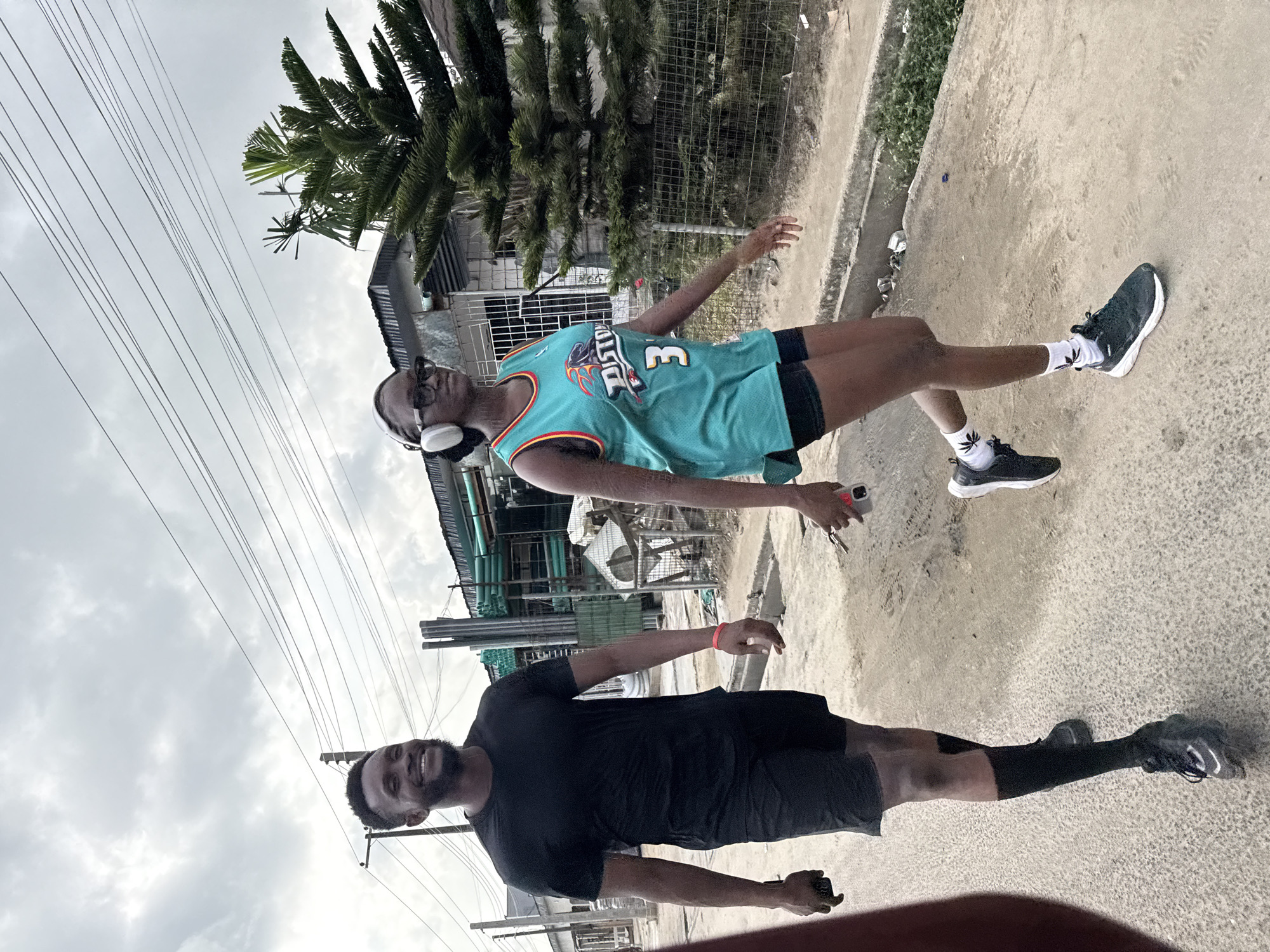 The image captures two young men outdoors in Ado, Nigeria, on what appears to be an overcast day. They are standing on a light-colored paved or concrete surface, possibly a road or path.
In the foreground, a man with a beard is smiling towards the camera. He is dressed in a black t-shirt, black shorts, black socks, and black sneakers. A red wristband is visible on his left wrist, and he holds a small, dark object in his left hand.
Slightly behind him and to the left, another man is posing. He wears a teal basketball jersey that displays the word "PISTON" and the number "3" on the front. He is also wearing black shorts, white over-ear headphones, and glasses. His white socks feature a repeating dark plant leaf pattern, and he wears black sneakers with white soles. He holds a light-colored smartphone in his right hand.
The background features various elements of an urban or semi-urban environment, including buildings with corrugated roofs, fences, and several trees, most prominently a tall, slender, evergreen-like tree. Multiple power lines crisscross the light grey, overcast sky.