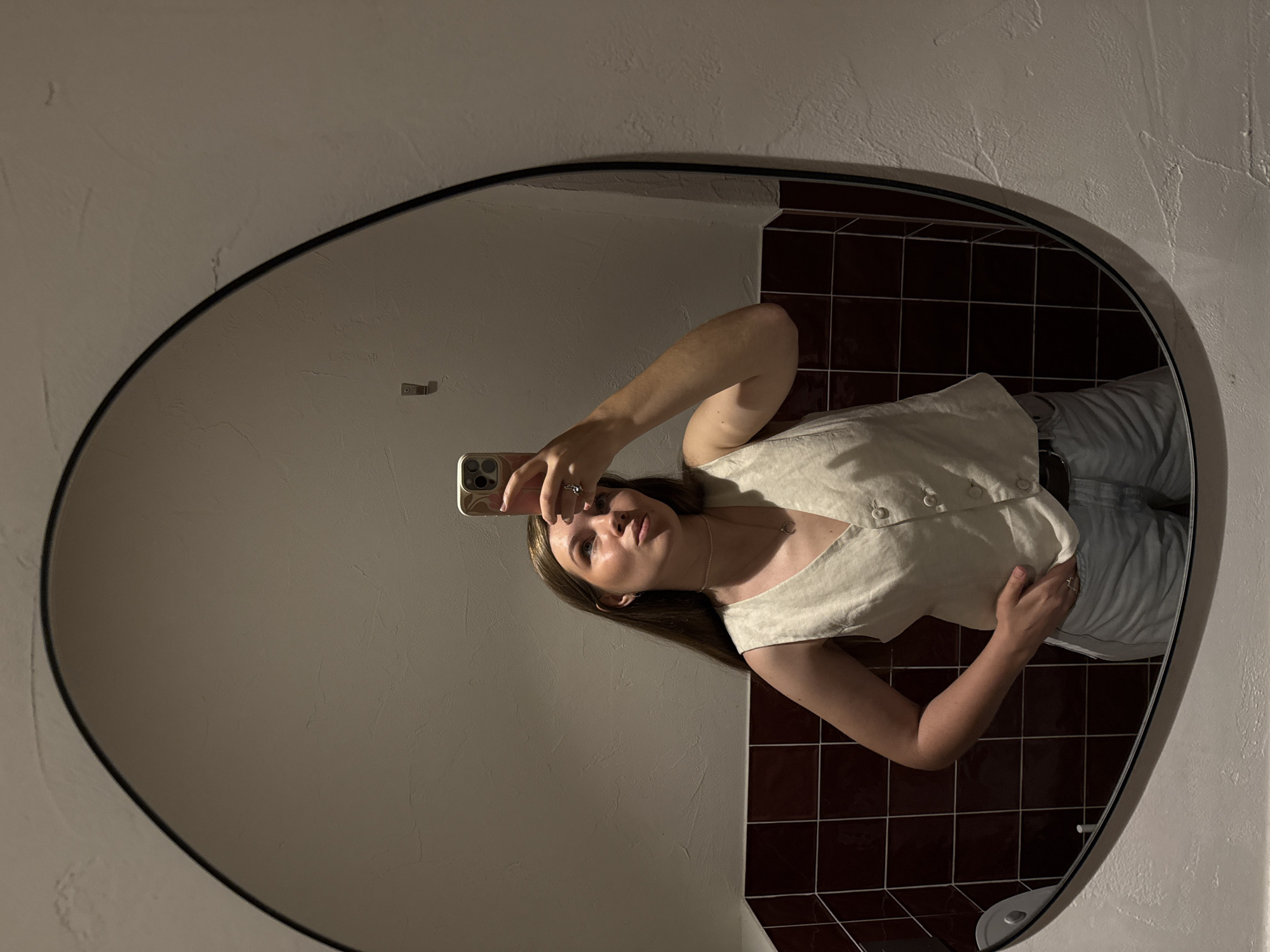 A woman is taking a selfie in an oval-shaped mirror. The woman has long brown hair and is wearing a white top and light-colored pants. She is holding her phone up with her right hand. The background behind her consists of two wall surfaces: one that is a dark, reddish-brown tiled wall and another that is light-colored. A towel holder is visible near the tiled wall. Based on the information provided, this location may be a bathroom in Belgrade, Serbia.