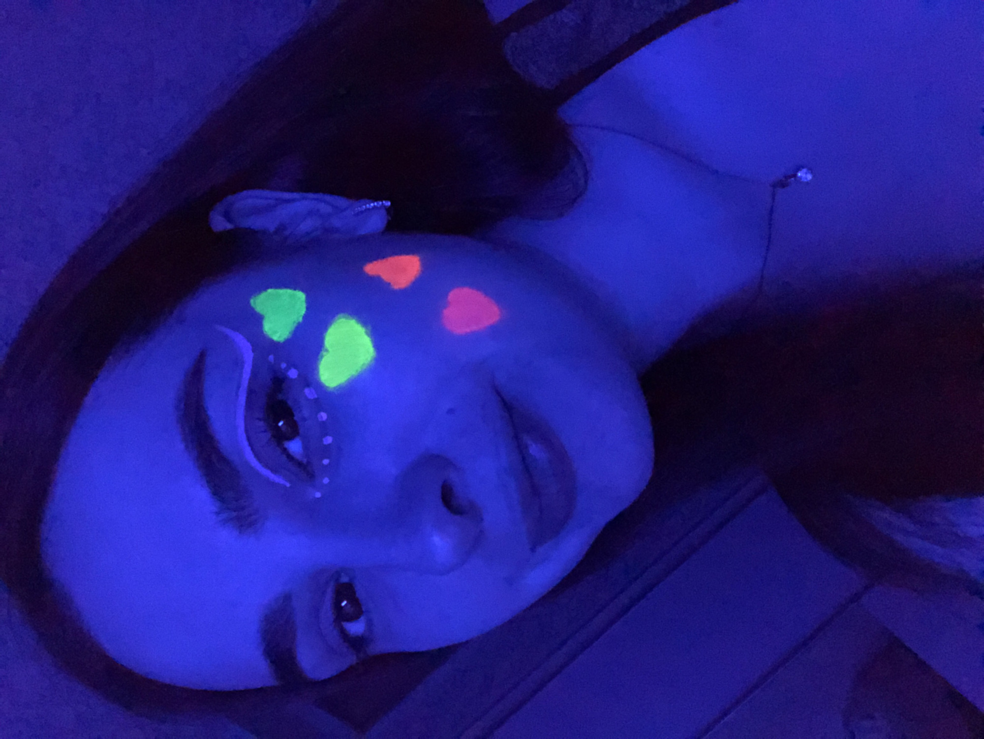 An angled, close-up shot captures a person, likely female, lying down and gazing towards the camera in a room lit by blacklight in Noviy, Russia. The dominant colors in the image are deep blue and purple, cast by the UV lighting, which makes certain elements glow.
The subject has long, dark hair visible along the top and left of the frame. Her face is prominently featured, adorned with striking fluorescent makeup. On her forehead and temple, several glowing heart shapes are visible: two bright green and two vivid orange/pink. Her right eye (from the viewer's perspective) is outlined with a bold, white or light blue glow-in-the-dark winged eyeliner, further accentuated by a series of small, glowing white dots applied beneath the lower lash line, extending outwards. Her dark eyebrows are well-defined, and her eyes are open. Her lips are slightly parted, and she maintains a neutral to subtly smiling expression.
She appears to be wearing a light-colored top, which absorbs the blue light, and a delicate necklace with a small, reflective pendant is visible on her chest. The background is dark and indistinct due to the low light conditions, suggesting an indoor setting. The blacklight effect implies the photograph was taken during the evening or night. There is no visible text in the image.