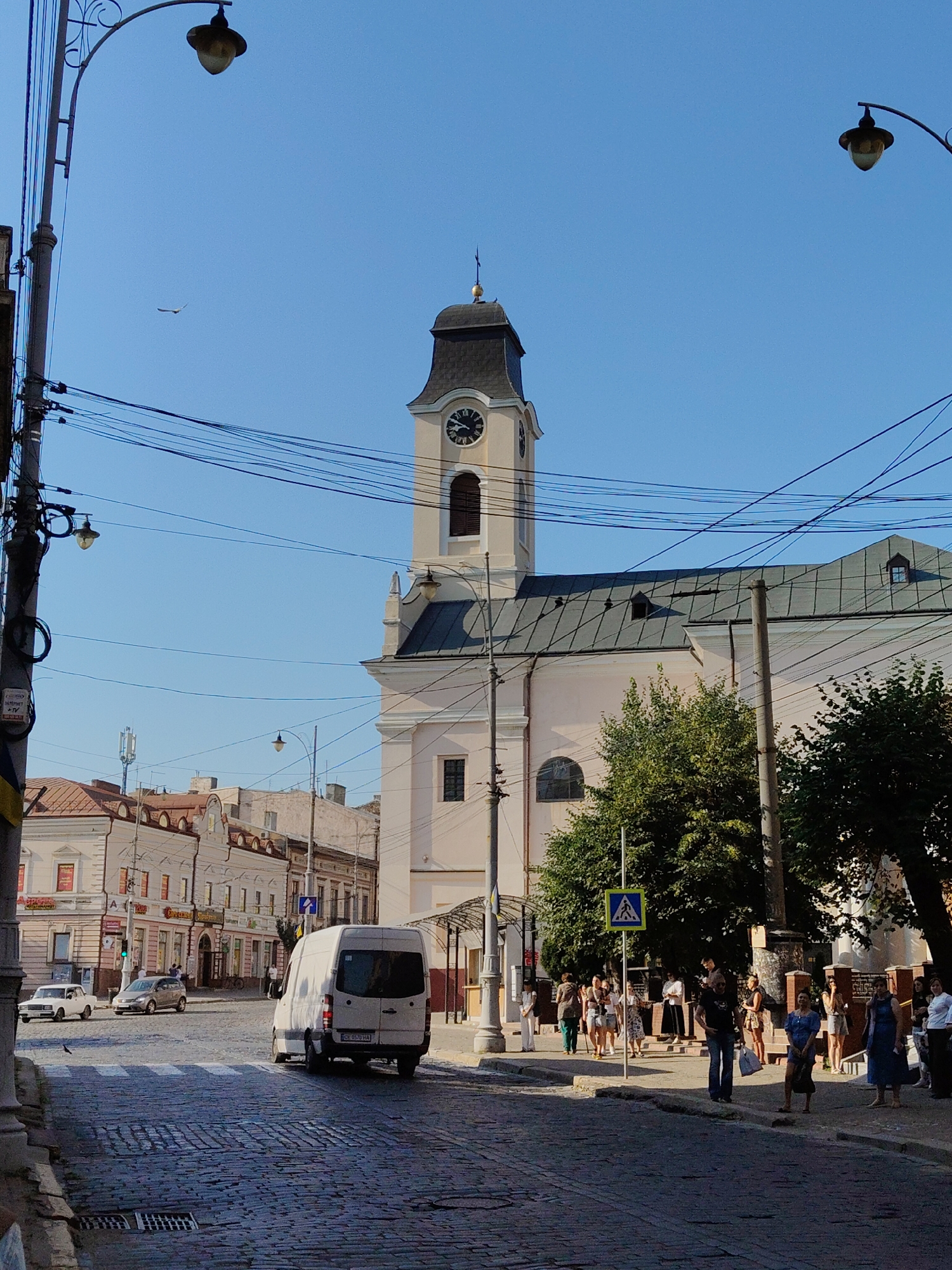 The image depicts a street scene in Chernivtsi, Ukraine, likely near the Chernivtsi National University, a UNESCO World Heritage Site. The focal point is a prominent clock tower belonging to a building that appears to be an educational institution or a historical landmark. The tower has a dark, multi-tiered roof and a clock face visible.
In the foreground, a cobblestone street curves, with a white cargo van driving away from the viewer. To the left, a white passenger car and a silver hatchback are parked or moving slowly. A pedestrian crossing is marked on the road.
On the right side of the street, a group of people are gathered on the sidewalk. They appear to be waiting or socializing. Some are standing, while others are sitting. The people are dressed in casual summer clothing. The background features buildings with varied architectural styles, some featuring arches and decorative elements. Trees with green foliage are interspersed throughout the scene. Overhead, a complex network of electrical wires and a street lamp are visible, typical of an urban environment. The sky is clear and blue. A bird is in flight in the upper left quadrant of the image.