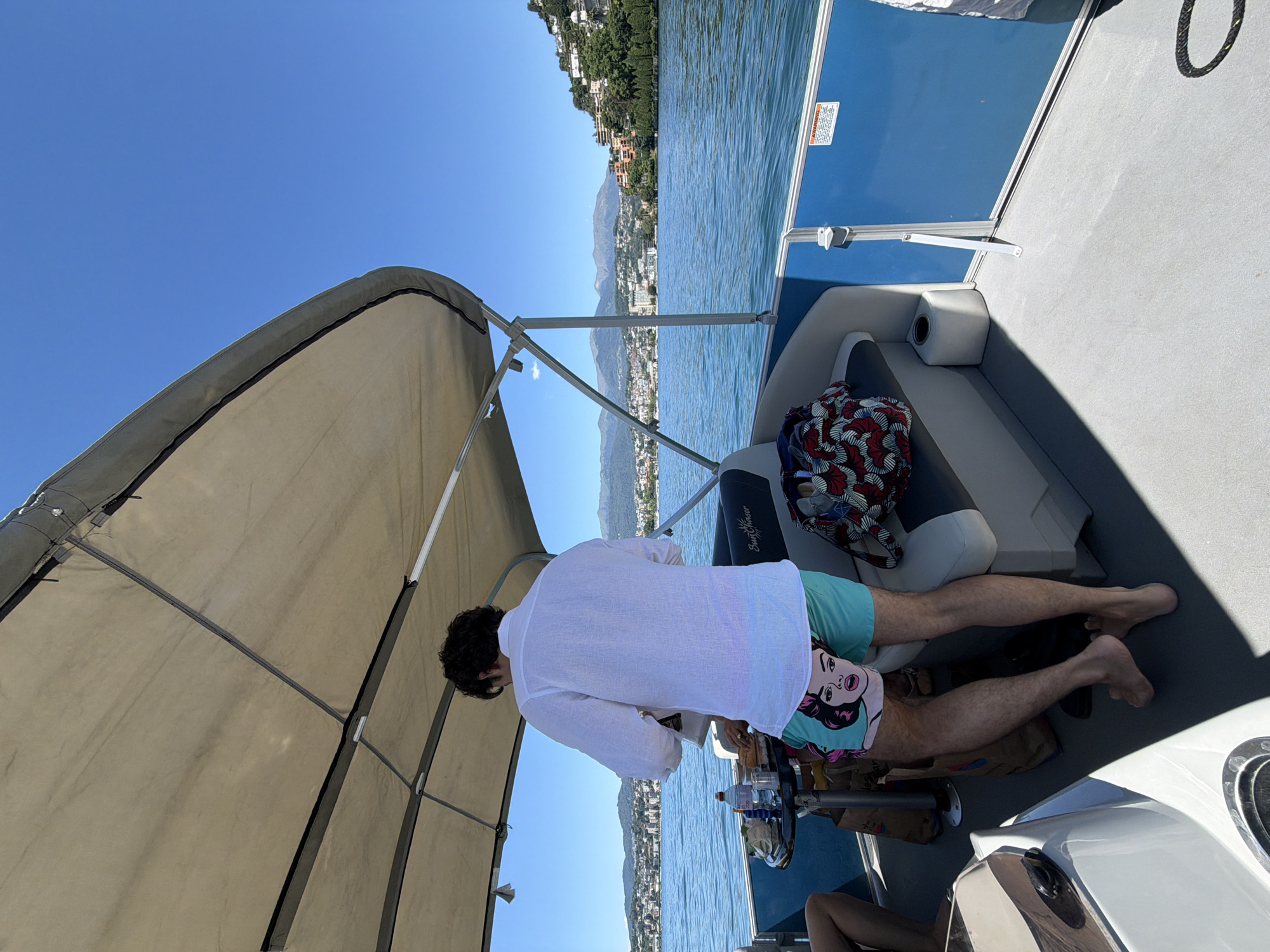 A man wearing a white linen shirt and teal swim trunks with a cartoon image on them stands on a pontoon boat on Lake Lugano in Switzerland. He is facing away from the camera and appears to be preparing food on a small table. A bottle of water and other items are on the table. The boat has a tan canopy and blue railing. In the background, the city of Lugano is visible on the shore, with buildings and mountains in the distance. The water is blue and calm.