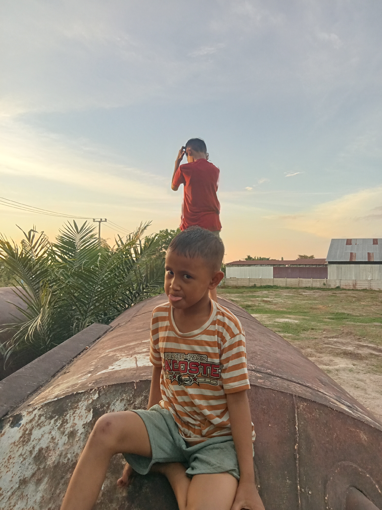 Two young boys are positioned outdoors on what appears to be a large, rusted metal structure. The boy in the foreground is seated, looking directly at the camera with his tongue sticking out slightly. He is wearing an orange and white striped t-shirt and light green shorts. The second boy is standing behind him, further up the structure, with his back to the camera, his right hand touching his head. He is wearing a red t-shirt.
Behind the boys, palm trees and other foliage are visible, along with a utility pole and power lines. In the background, several buildings with corrugated metal roofs and white walls can be seen, alongside a concrete barrier. The ground is covered with dry grass and dirt. The sky is clear with a gradient of blue and orange hues, suggesting it is late afternoon or early evening. The overall scene suggests an informal, possibly rural or semi-urban, outdoor environment in Kualu, Indonesia.