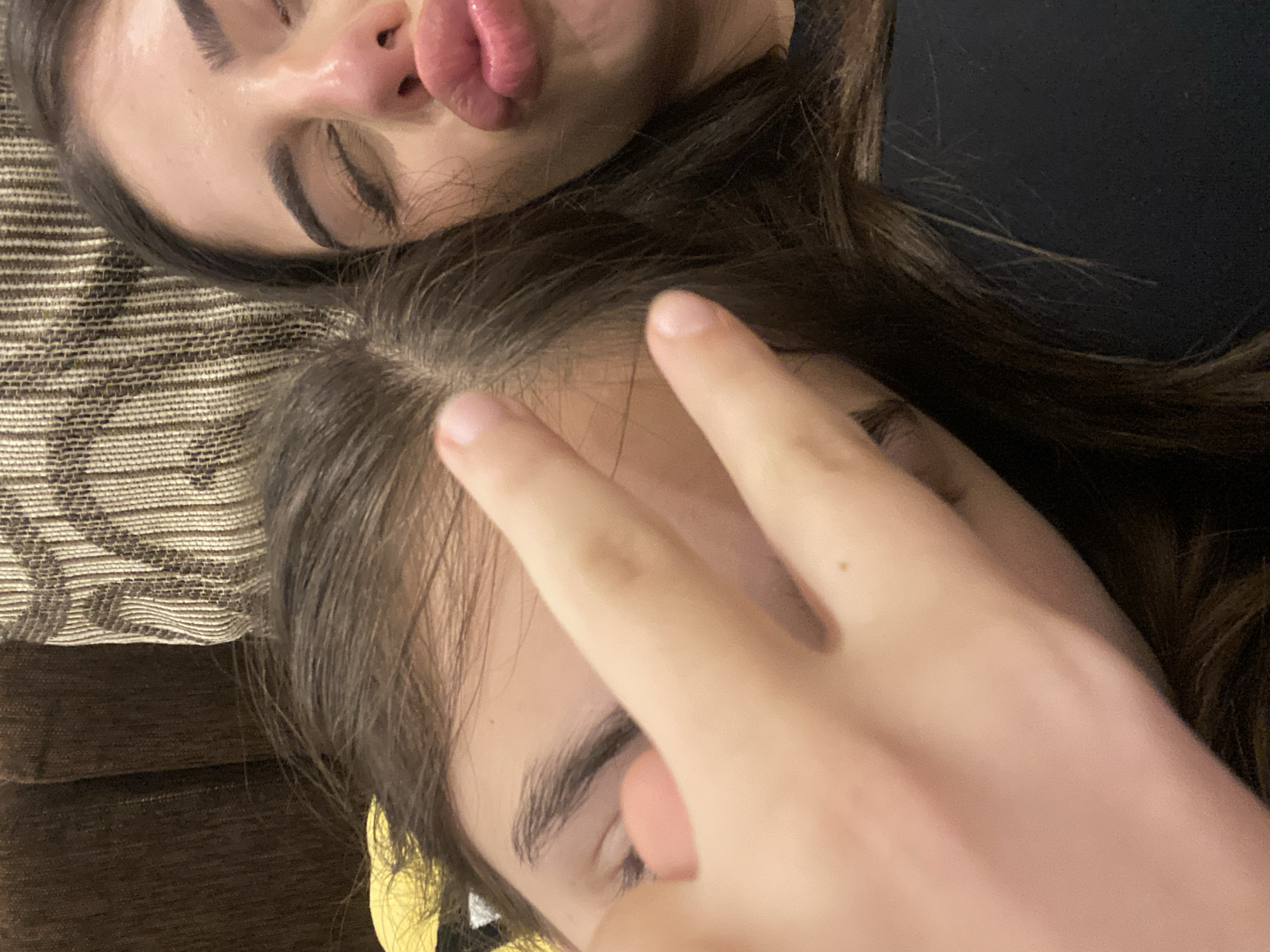 The image shows two people lying down next to each other. The person on the top has dark hair, closed eyes, and pursed lips. The person below has dark hair and is making a peace sign with their fingers covering part of their face. The background includes a textured cushion or couch. There are no indications of the city of Dragovishtitsa, Bulgaria.
