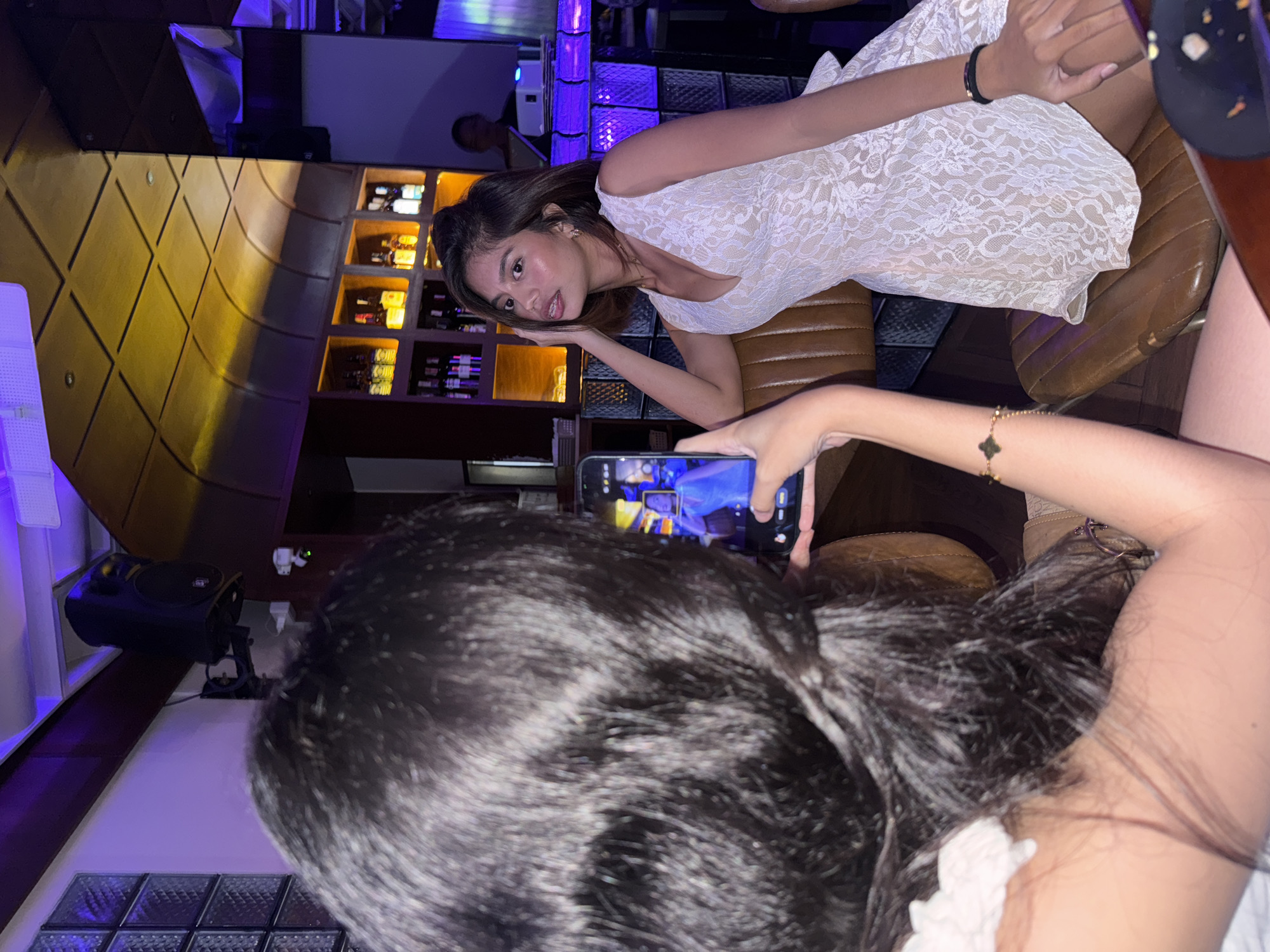 The image depicts two young women indoors, likely at a bar or restaurant in Davao City, Philippines, given the context. One woman, with dark hair covering much of her face, is holding up a smartphone to take a picture of the other woman. The second woman, seated and facing the camera, has short, dark hair and is wearing a white lace dress. She is looking away from the camera, with her left arm resting on her head and her right hand on her thigh. The background reveals a bar area with shelves stocked with bottles, illuminated by warm, golden lighting, and accented with purple and blue lights, suggesting a lively evening atmosphere. The woman on the left appears to be the one taking the photo, and her arm with a bracelet is visible holding the phone. The setting suggests a social outing.