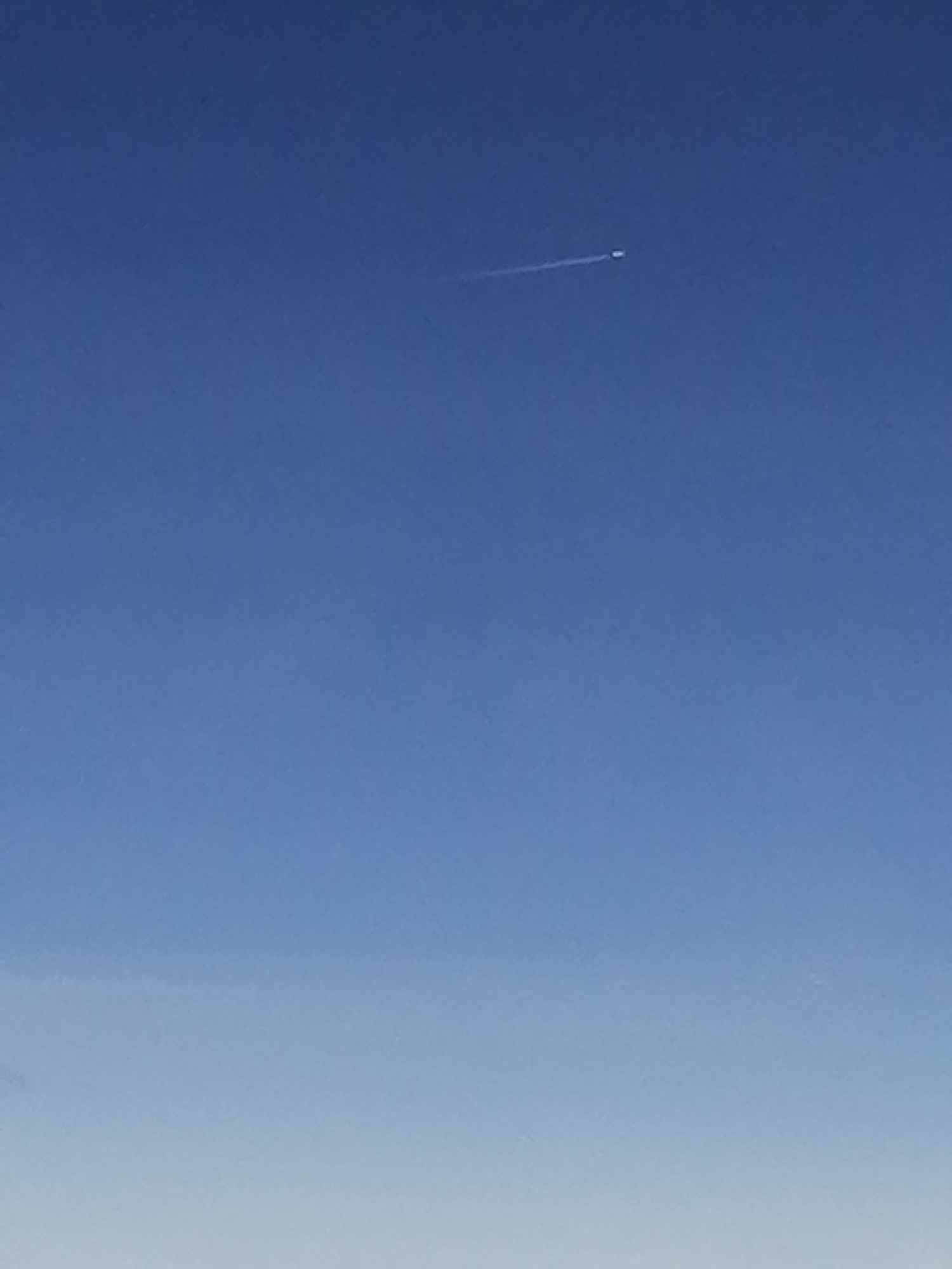 The image shows a clear blue sky. A distant airplane is visible in the upper portion of the frame, leaving a white contrail. The lower portion of the image displays a lighter shade of blue, possibly indicating the horizon. The location is Unknown, Unknown.