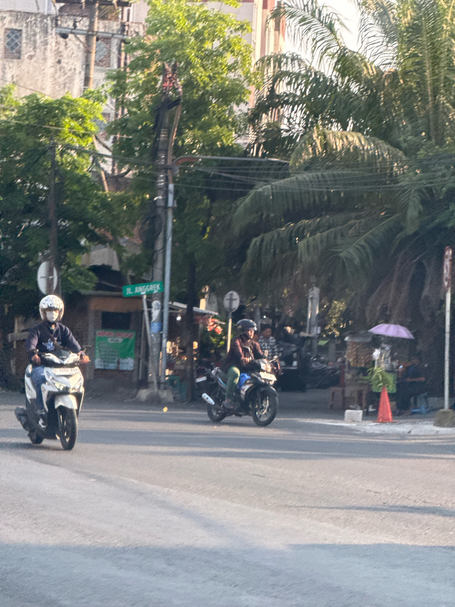 The image captures a street scene in Surabaya, Indonesia, likely during the late afternoon due to the warm sunlight casting long shadows. In the foreground, a man wearing a white helmet and a dark top is riding a white scooter on a paved road. Further down the road, another person, also wearing a helmet, is riding a motorcycle. The background reveals a mix of urban elements. A tall building with windows is visible on the left, interspersed with lush green trees. A street sign with "JL. ANGGREK" written in green is mounted on a utility pole. Several other motorcycles and people are present in the mid-ground, some appearing to be at a roadside stall. A purple umbrella and an orange traffic cone are also visible. The overall environment suggests a busy, everyday street in a tropical city.