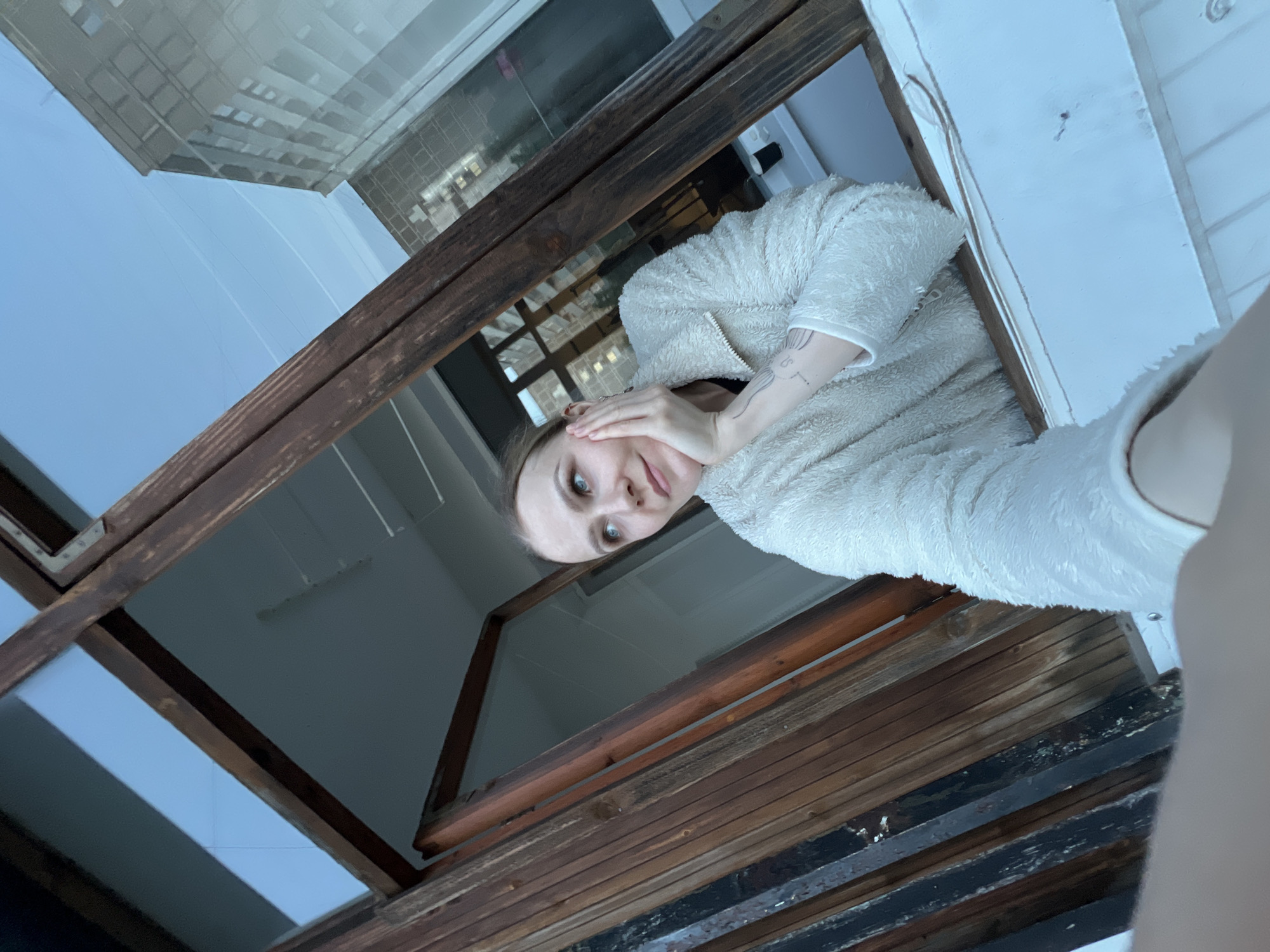 A young woman is depicted looking down through an opening, possibly a skylight or an attic window, framed by rustic wooden beams. She is positioned in the upper right portion of the image, gazing directly towards the viewer with a neutral to slightly pensive expression. Her left hand is raised to her face, resting on her cheek, while her right hand is partially obscured. She is dressed in a light-colored, fluffy jacket. A tattoo of delicate, abstract lines is visible on her left forearm.
Beyond the wooden frame and through the glass panes of the opening, a clear blue sky is visible, indicating daytime with fair weather. Reflections of multi-story, modern buildings with numerous windows can be seen in the glass, suggesting an urban environment. The setting for this scene is Khimki, Russia. No explicit text is visible in the image apart from the abstract lines of the tattoo.