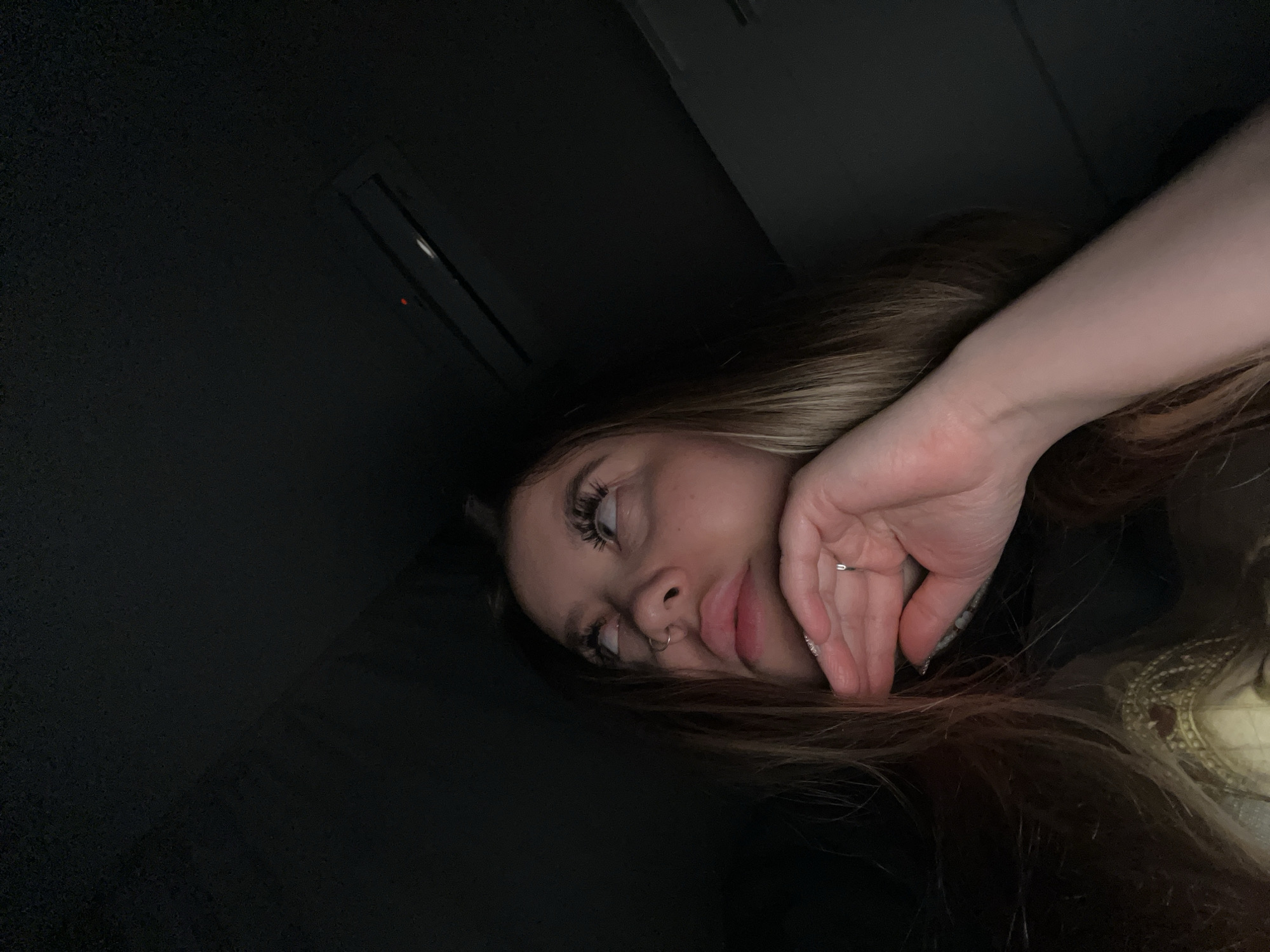 The image is a dimly lit, close-up portrait of a young woman, taken in Krasnodar, Russia. She is depicted from a slightly high angle, resting her head on her right hand, which bears a silver ring on its fourth finger. Her long, light brown hair frames her face. Notable features include prominent eyelashes and a silver hoop nose piercing. Her lips are slightly parted, and her gaze is directed upwards and to the left of the frame, conveying a contemplative or relaxed expression.
Part of a decorative garment or accessory, possibly embroidered in light colors with subtle red accents, is partially visible on her right side. The background is predominantly very dark, suggesting an indoor setting or nighttime conditions. In the upper left quadrant of the background, a rectangular panel with a small red indicator light can be faintly seen, which is consistent with the interior of a vehicle, such as an overhead console, or a similar room fixture. No text is visible in the image.