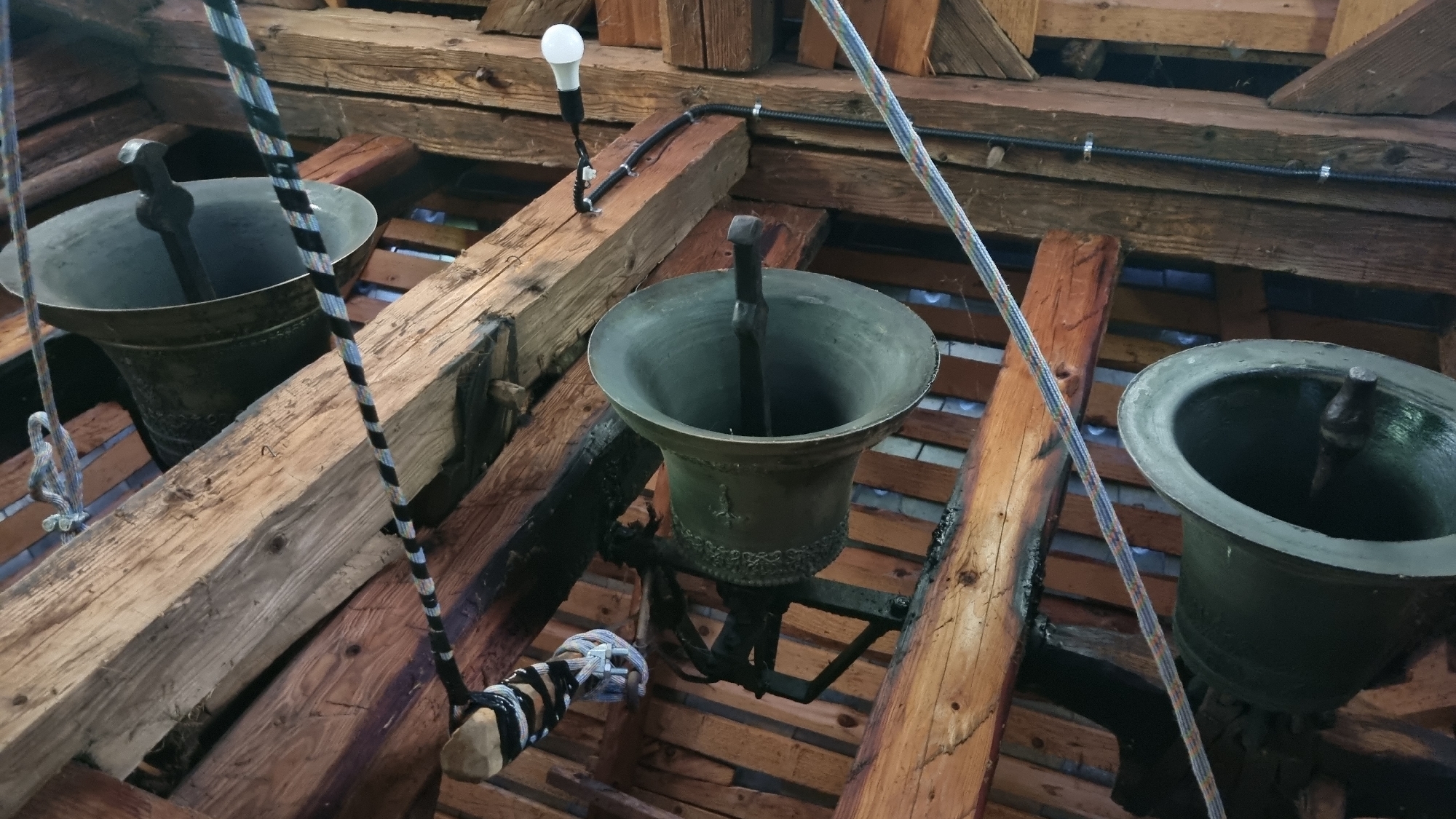 The image shows a close-up view of several large, dark-colored bells suspended from wooden beams in what appears to be a belfry. The bells are made of metal and have a classic bell shape, with clappers visible inside. Ropes are attached to the bells, suggesting they are meant to be rung. The wooden structure is rustic and appears old, with visible grain and some charring on certain beams, possibly from age or past events.
In the background, a single white light bulb is illuminated, attached to a wire that runs along the beams. There are also electrical cables and conduits visible, indicating that the belfry has been equipped with modern lighting. The overall scene is dimly lit, with light primarily coming from the single bulb and some ambient light filtering from above or through unseen openings. The perspective is from below, looking upwards at the bells and the wooden ceiling of the belfry.
The scene is static, with no human activity directly depicted, though the presence of ropes implies past or potential use for ringing the bells. The focus is on the bells themselves and the structural elements of the belfry. The setting suggests a historical or religious location, likely a church or bell tower, given the presence of bells. The condition of the wood and the rustic nature of the structure point towards an older building. No text is visible in the image. The location context provided is Vyshka, Ukraine.