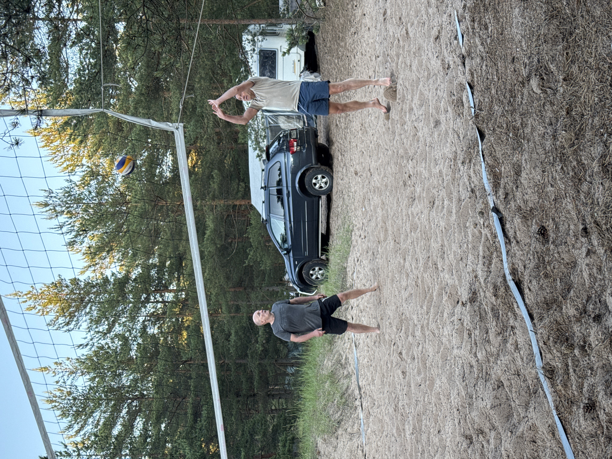 Two adult males are playing volleyball on a sandy court surrounded by pine trees. One male, wearing a gray t-shirt and black shorts, stands on the left side of the court, facing right. The other male, wearing a tan t-shirt and blue shorts, is in mid-air on the right side of the court, with his arms raised in a spiking motion. A volleyball is visible above the net between them. In the background, a dark colored SUV and a white camper trailer are parked. The scene appears to be in a recreational area near Ozerki (Krasnodolinskaya), Russia, given the pine forest environment.