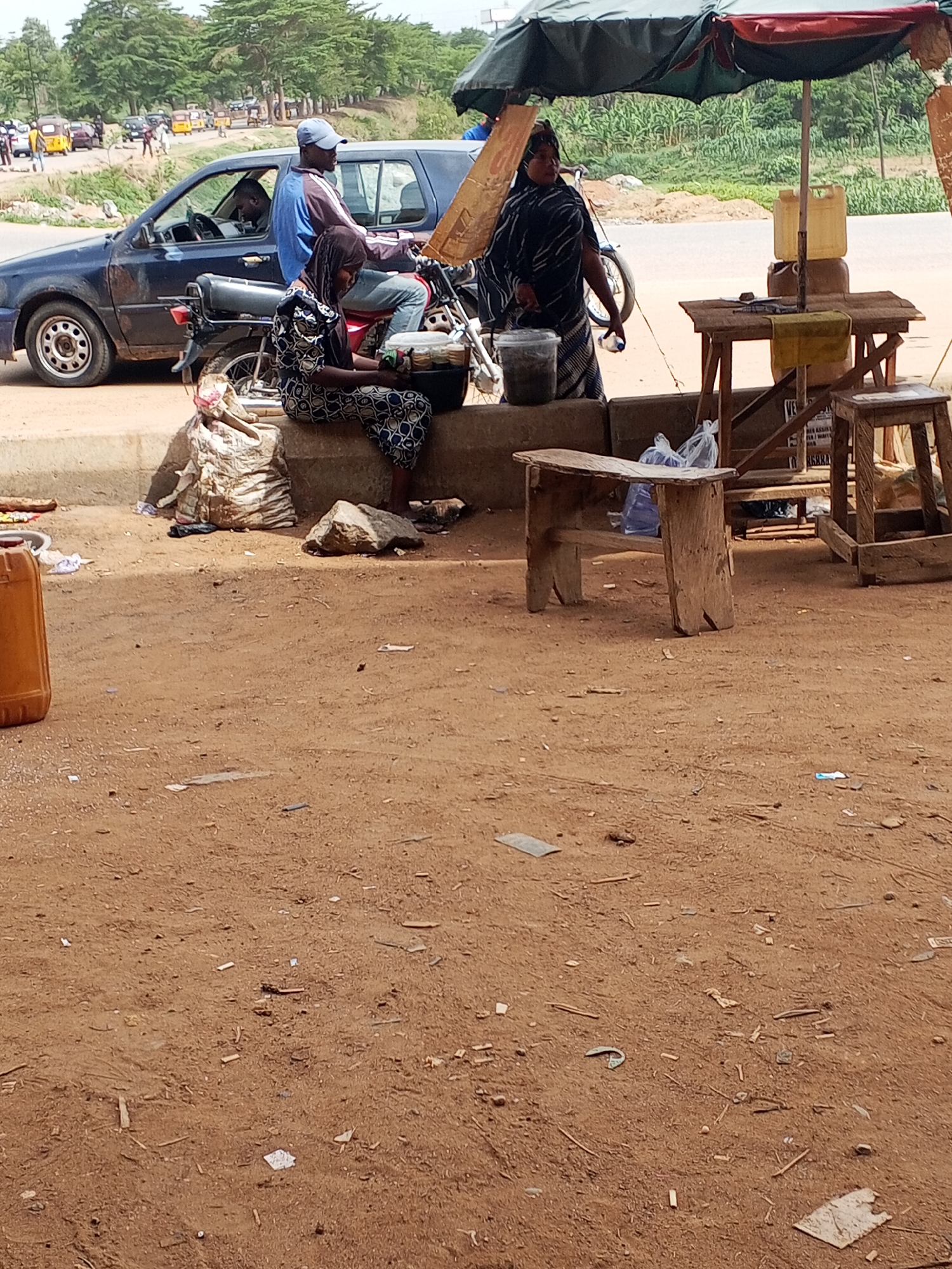 The scene takes place in Galadimawa Village, Nigeria. A woman in a patterned dress sits on a concrete barrier next to a motorcycle occupied by a man in a blue jacket and cap. Another woman in a striped garment stands under a green umbrella, next to a table with yellow containers and various items. A blue car is parked nearby. The foreground is dominated by a dirt ground scattered with debris. Wooden benches are also present. Traffic passes in the background.