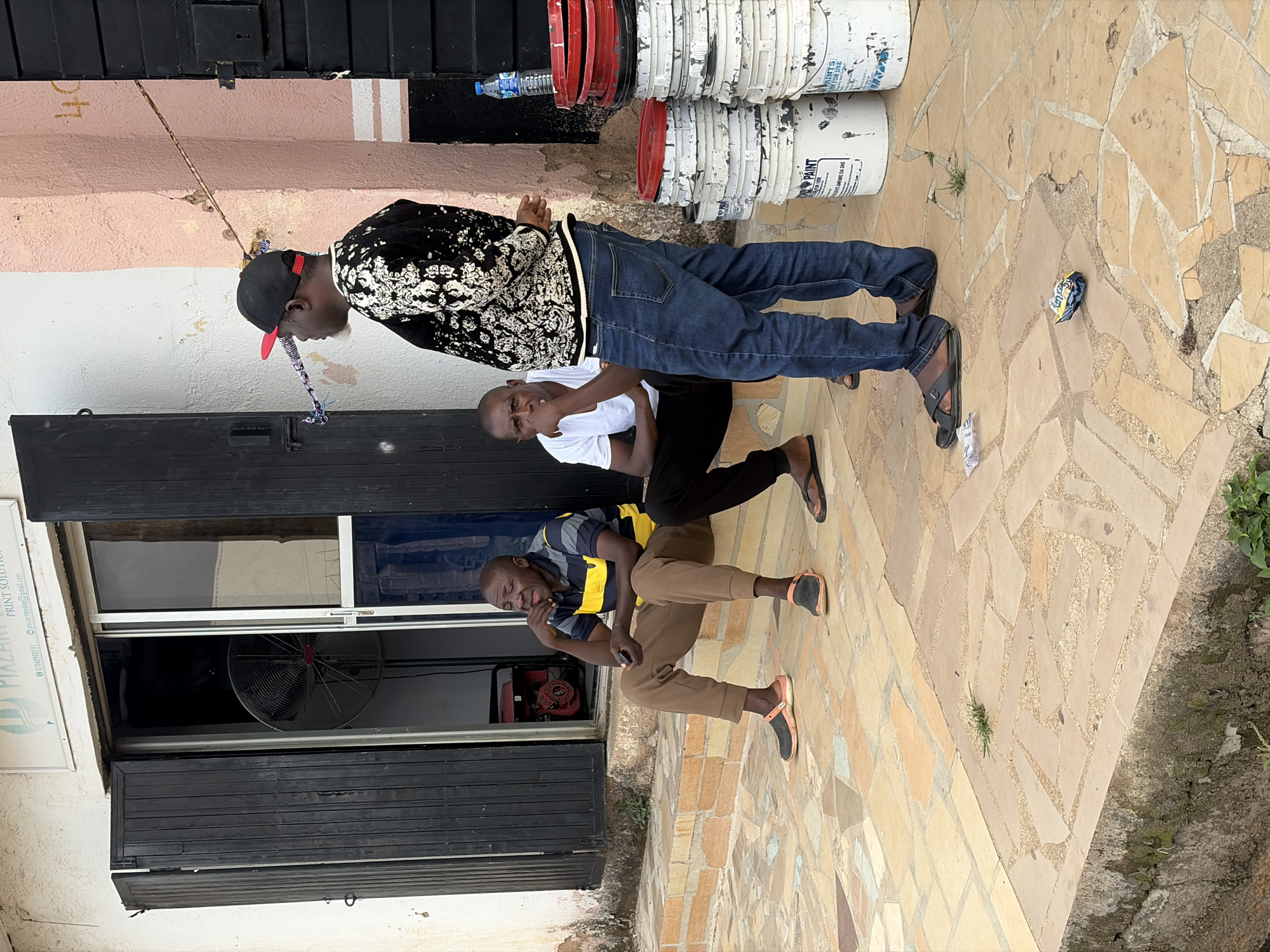 This image depicts a casual street scene in Jos, Nigeria, focusing on three adult Black men gathered outside a commercial establishment.
To the left, a middle-aged man sits on a low, tiled concrete step. He wears a horizontal striped polo shirt (dark blue, yellow, light blue), brown pants, and black slip-on shoes with orange accents. He is leaning slightly forward, with his right hand near his mouth or chin, and appears to be contemplating or listening. Adjacent to him, further to the right on the same step, another middle-aged man sits, wearing a white short-sleeved t-shirt, dark pants, and black flip-flops. His left hand is also near his chin, and he looks generally forward.
Standing to the right, with his back mostly to the viewer, is a third middle-aged man. He wears a black and white patterned long-sleeved top or sweater, blue denim jeans, and black flip-flops. A red and black baseball cap is worn backward on his head, and he has visible white/grey stubble on his chin. He stands in profile, facing left, seemingly observing or participating in a conversation with the other two men. A decorative, wrapped string or fabric object is visible tied between two points on the wall behind him.
The background shows the exterior of a building with a white facade. A sign partially visible above the entrance reads "PIXZART PRINT SOLUTION" with contact details. The entrance features dark, folded wooden or metal shutters partially open, revealing a sliding glass door behind them. Inside the shop, a large black standing fan and a red and black portable generator are visible. To the far right, a pale pink structural pillar or wall section shows the faint number "40" painted on it, indicating an address.
Stacked against the right wall are approximately nine white paint buckets with red lids, some bearing the label "DURAPAINTS FOR USE." A plastic water bottle with a blue cap rests on top of the stack.
The ground in the foreground consists of irregularly shaped, multi-toned flagstone-style paving in shades of beige, tan, and light brown. A small crumpled blue and yellow wrapper and a piece of white paper litter the ground near the standing man's feet. Small tufts of grass and moss grow in cracks in the paving and along the base of the building's facade.