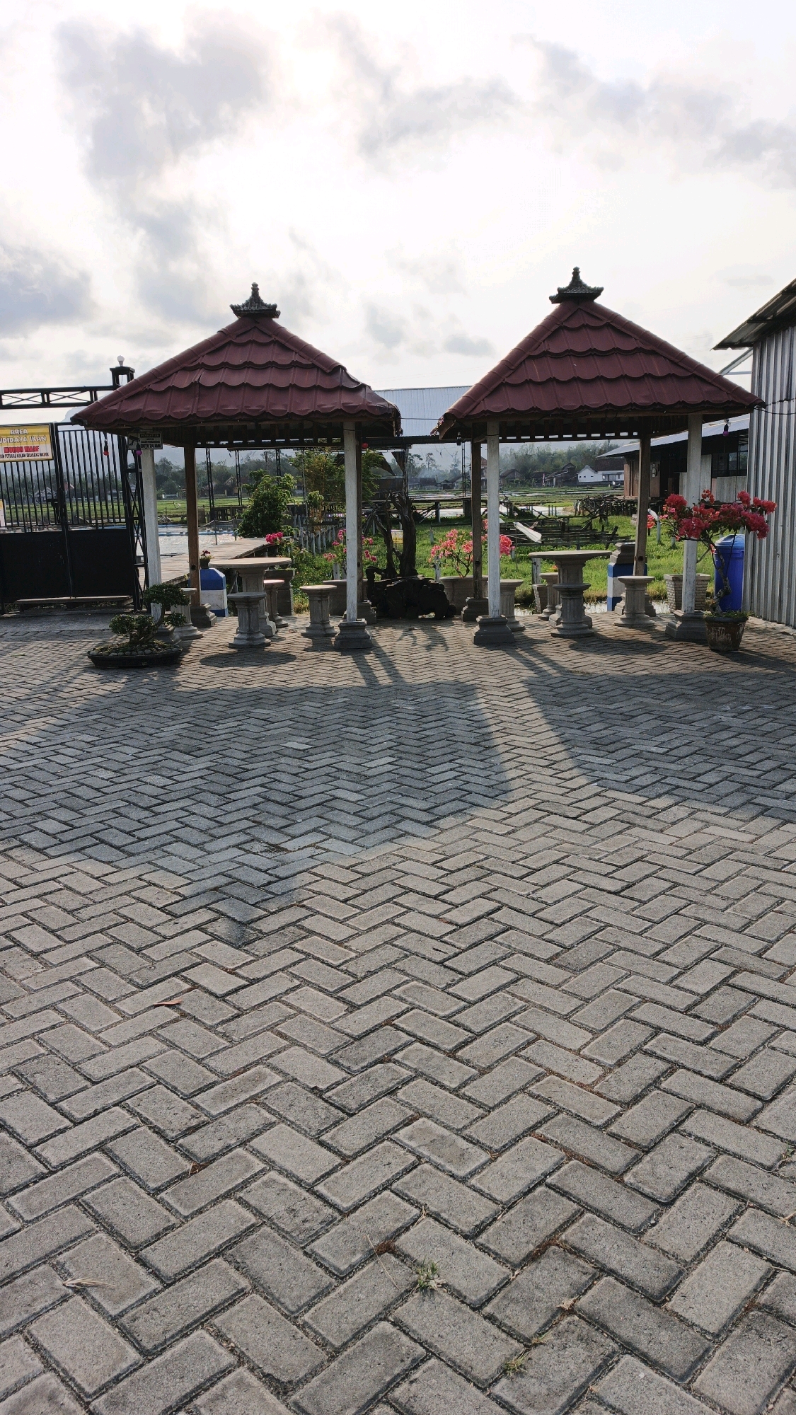 The image depicts a paved outdoor area featuring two red-tiled, open-sided gazebos. Each gazebo has a central table with attached seating arrangements, appearing to be made of stone. In front of the gazebos, the ground is paved with interlocking gray bricks laid in a herringbone pattern. A black metal gate is visible to the left, with a sign attached to it. Behind the gazebos, a green landscape with trees and some structures can be seen. To the right, a building with a corrugated metal exterior is present. The scene appears to be an outdoor seating area, possibly in a park or a recreational facility, and is located in Tanggulwelahan, Indonesia. The lighting suggests it is daytime, with shadows cast by the gazebos and their supports on the brick pavement. There are no people visible in the image.